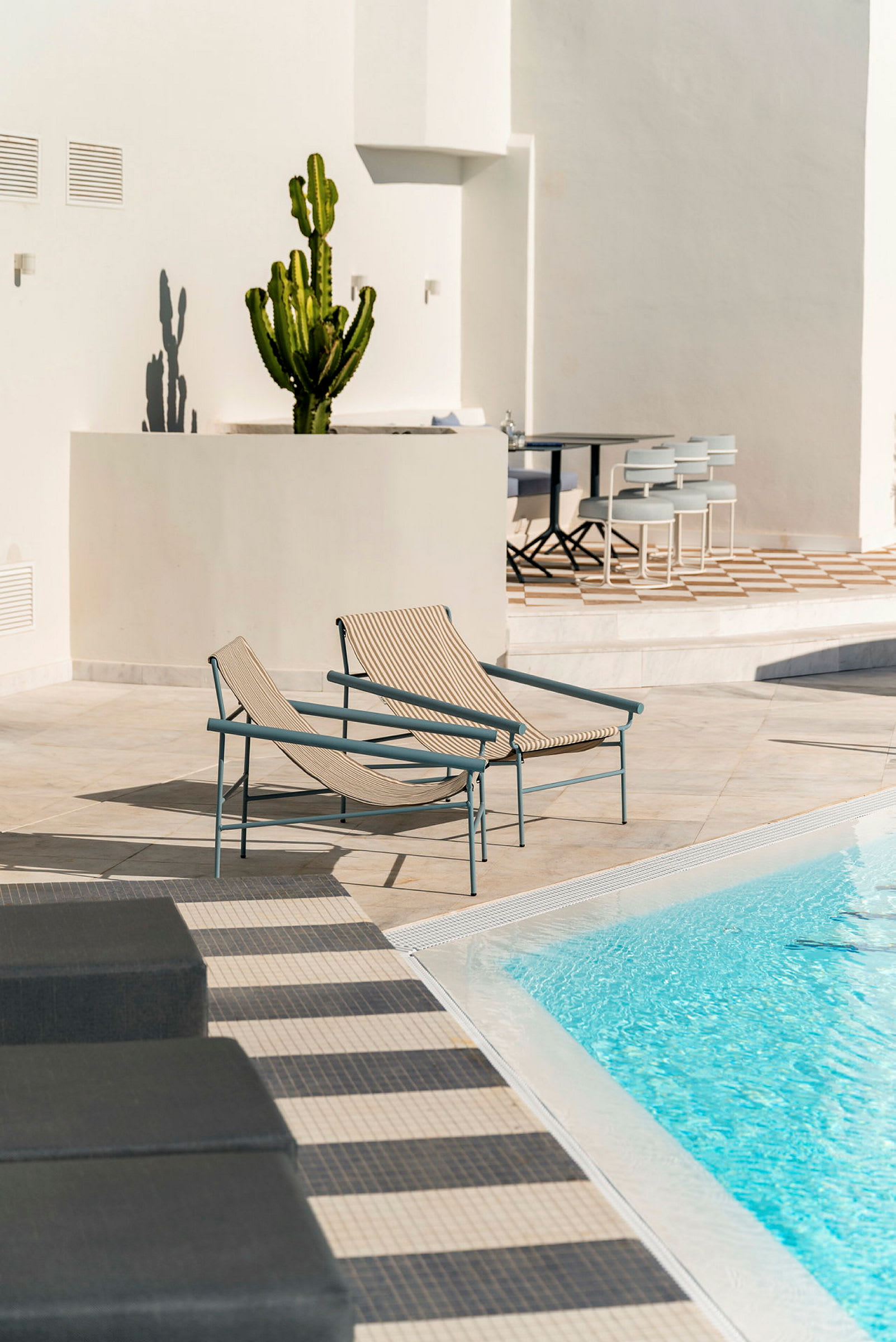 Två solstolar nära en pool med klarblått vatten. Bakgrunden visar en vit vägg och en kaktus samt en matplats. (AI-genererad text)