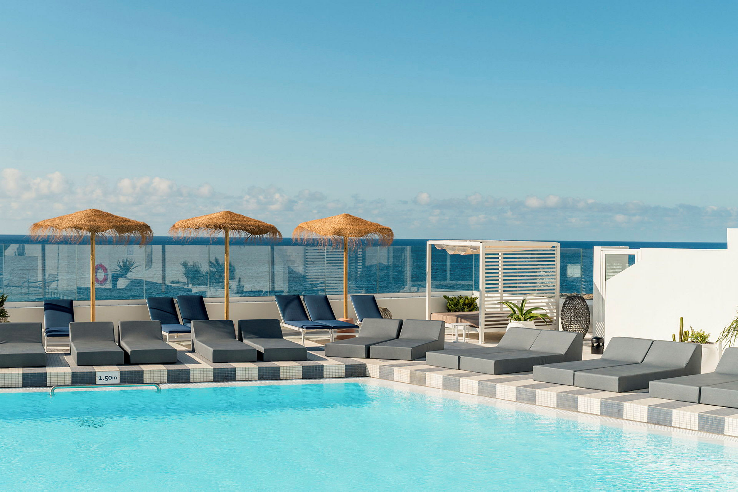 En pool med solstolar och parasoller av halm, med havet i bakgrunden. (AI-genererad text)