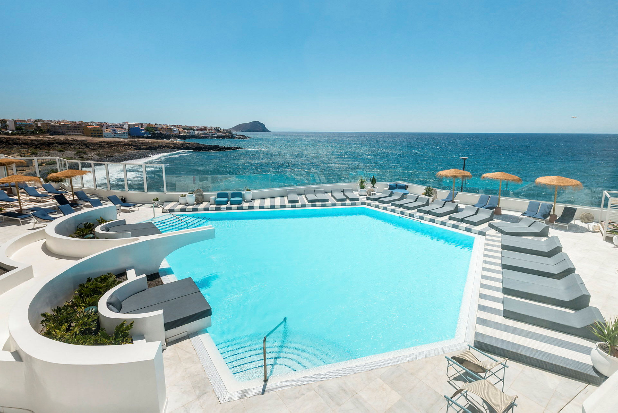 En modern pool med solsängar och parasoller med utsikt över havet och en kustlinje i bakgrunden. (AI-genererad text)