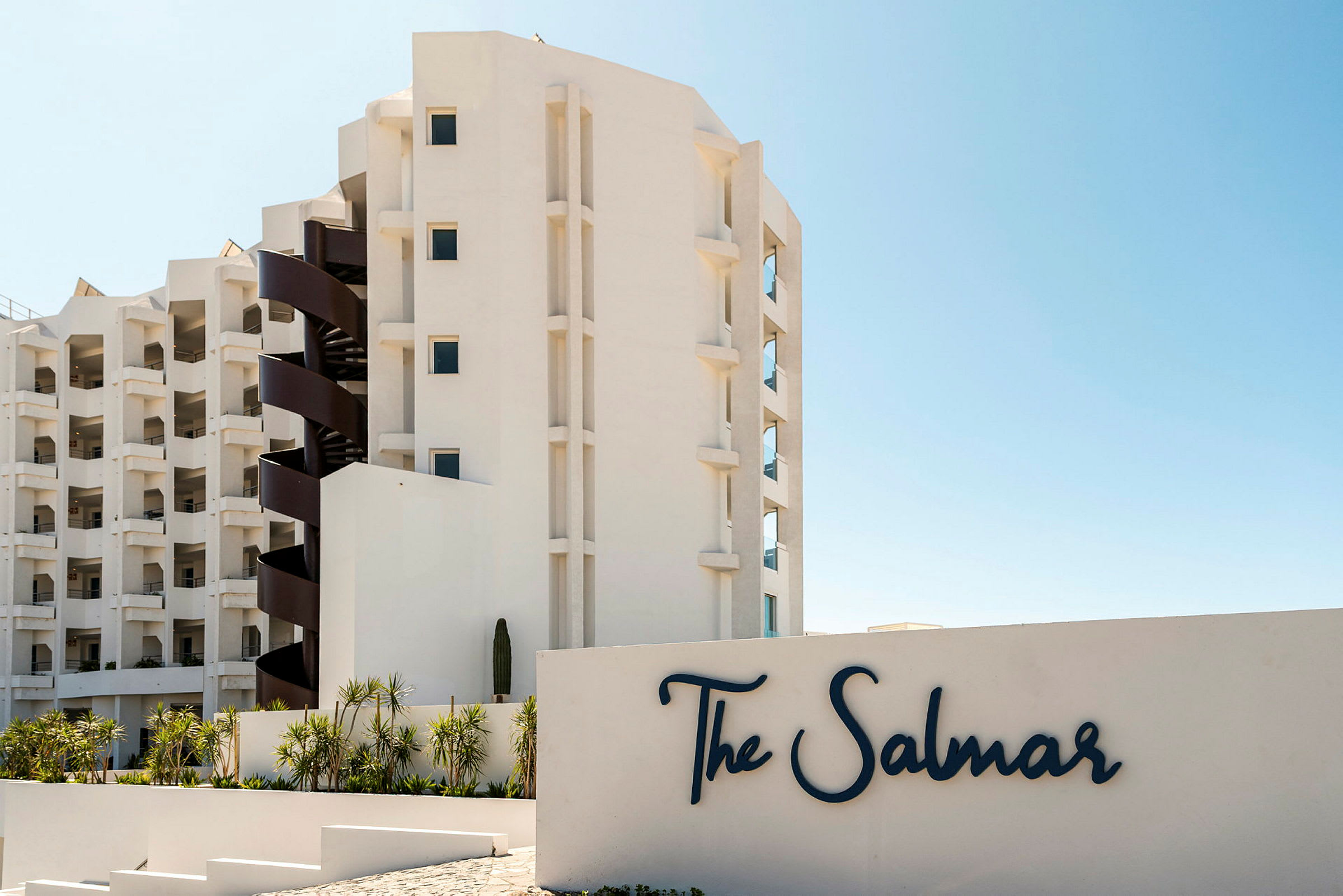 En modern byggnad med balkonger och en spiraltrappa bredvid en skylt med texten "The Salmar". Framför byggnaden finns planteringar med palmer. (AI-genererad text)
