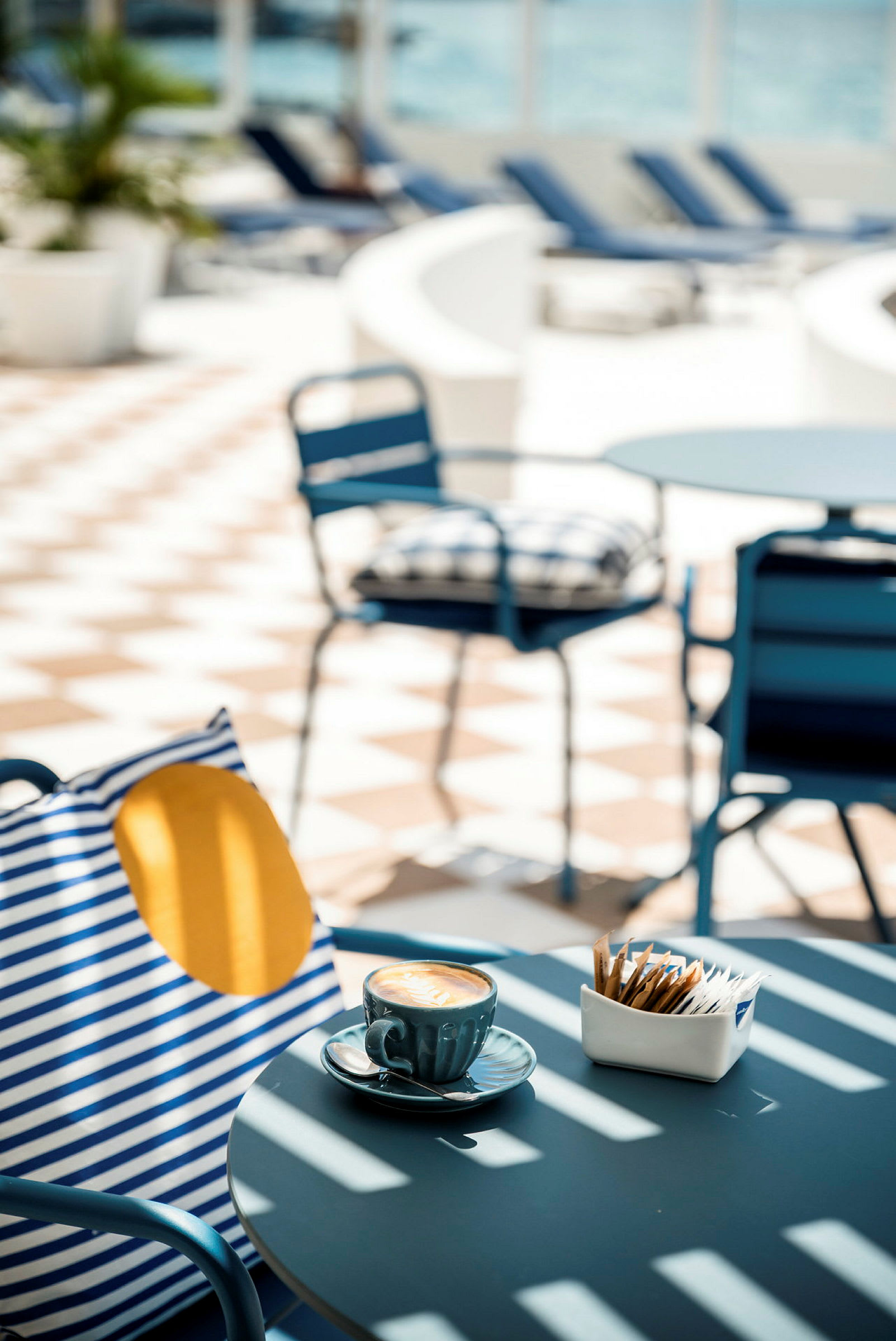 Ett bord med en kopp kaffe och sockerpaket står i ett soldränkt utomhusområde med möbler. (AI-genererad text)