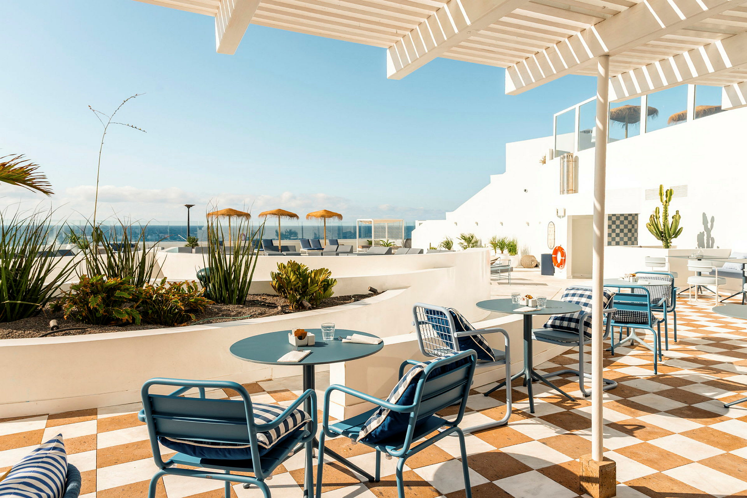 Uteservering med bord och stolar under en pergola, omgiven av grönska och med utsikt över havet. (AI-genererad text)