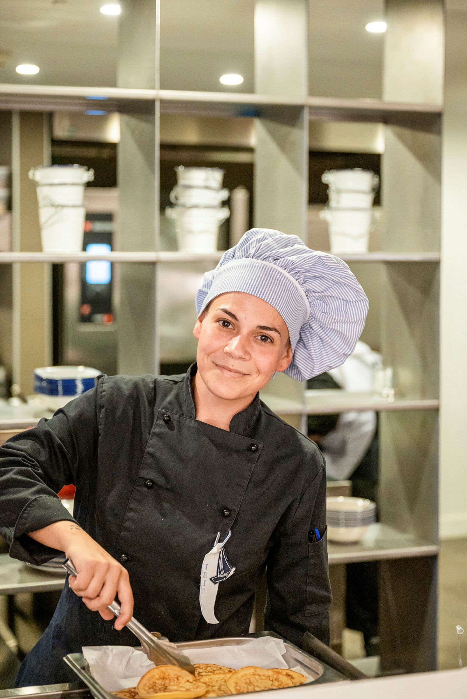 En kock i arbetskläder plockar upp pannkakor från en plåt med hjälp av en tång i ett kök. (AI-genererad text)