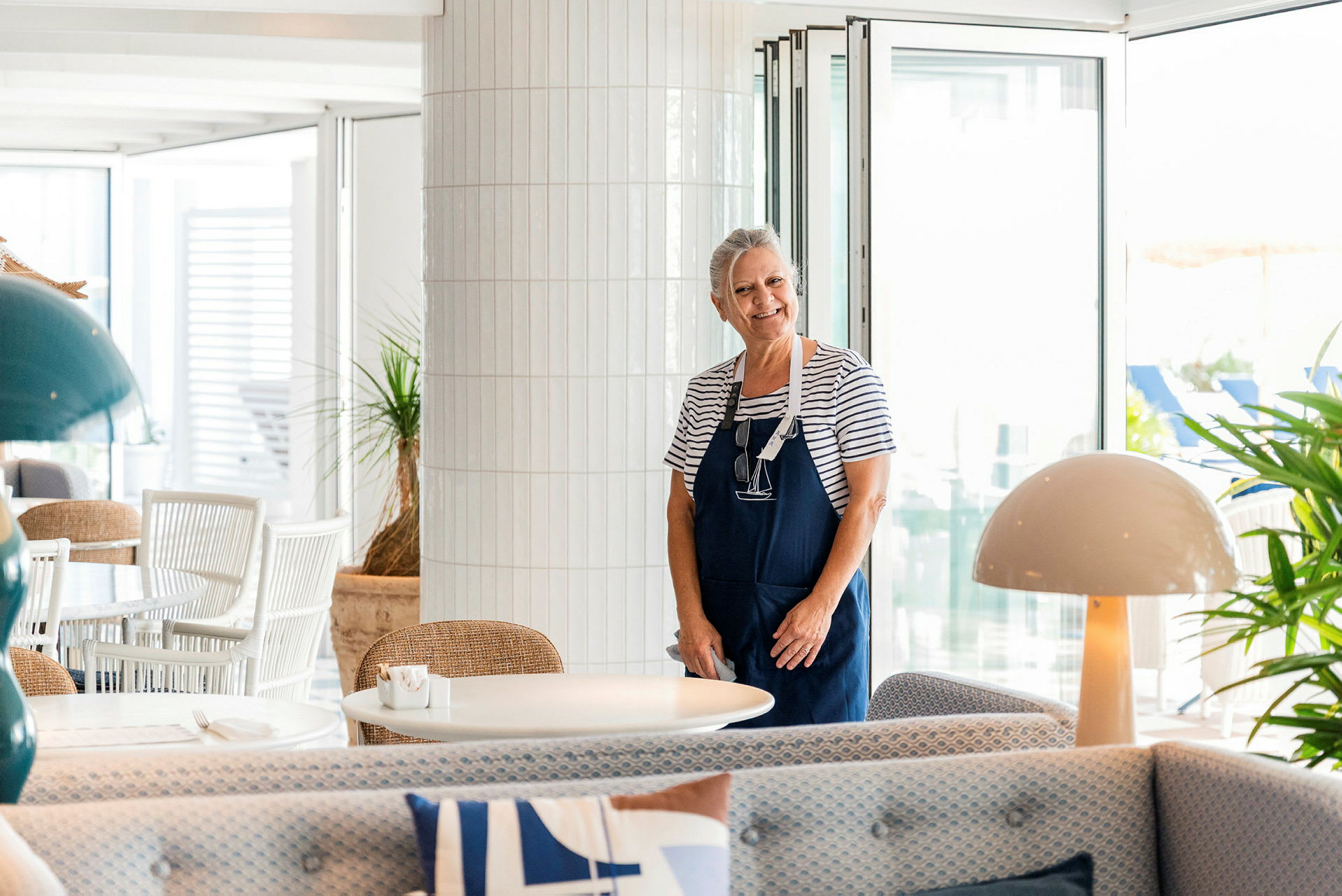 En kvinna står i en ljus och modern restaurang, klädd i randig tröja och förkläde. (AI-genererad text)