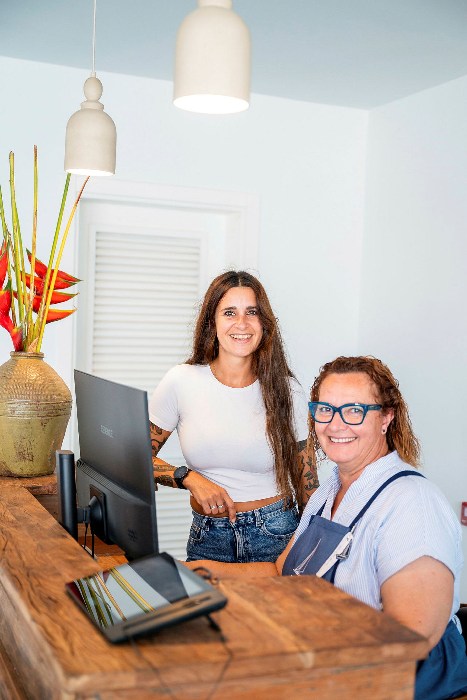Två personer står vid en receptionsdisk med en dator och en vas med röda blommor. (AI-genererad text)