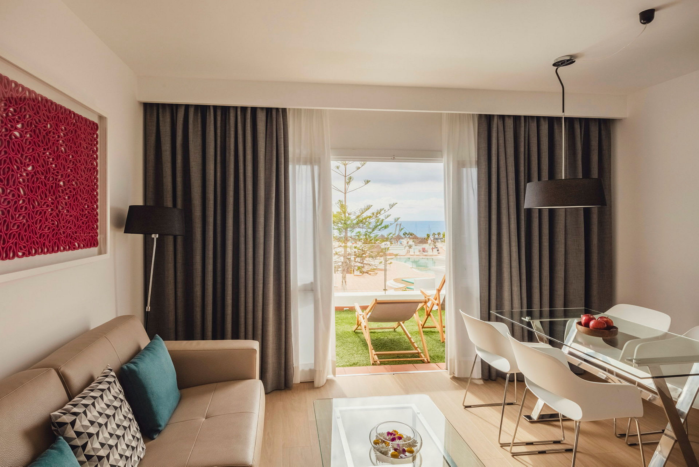 Ett modernt vardagsrum med en beige soffa och ett matbord med vita stolar. Rummet har stora fönster med gardiner och en utsikt mot en grön uteplats med solstolar samt en pool. (AI-genererad text)