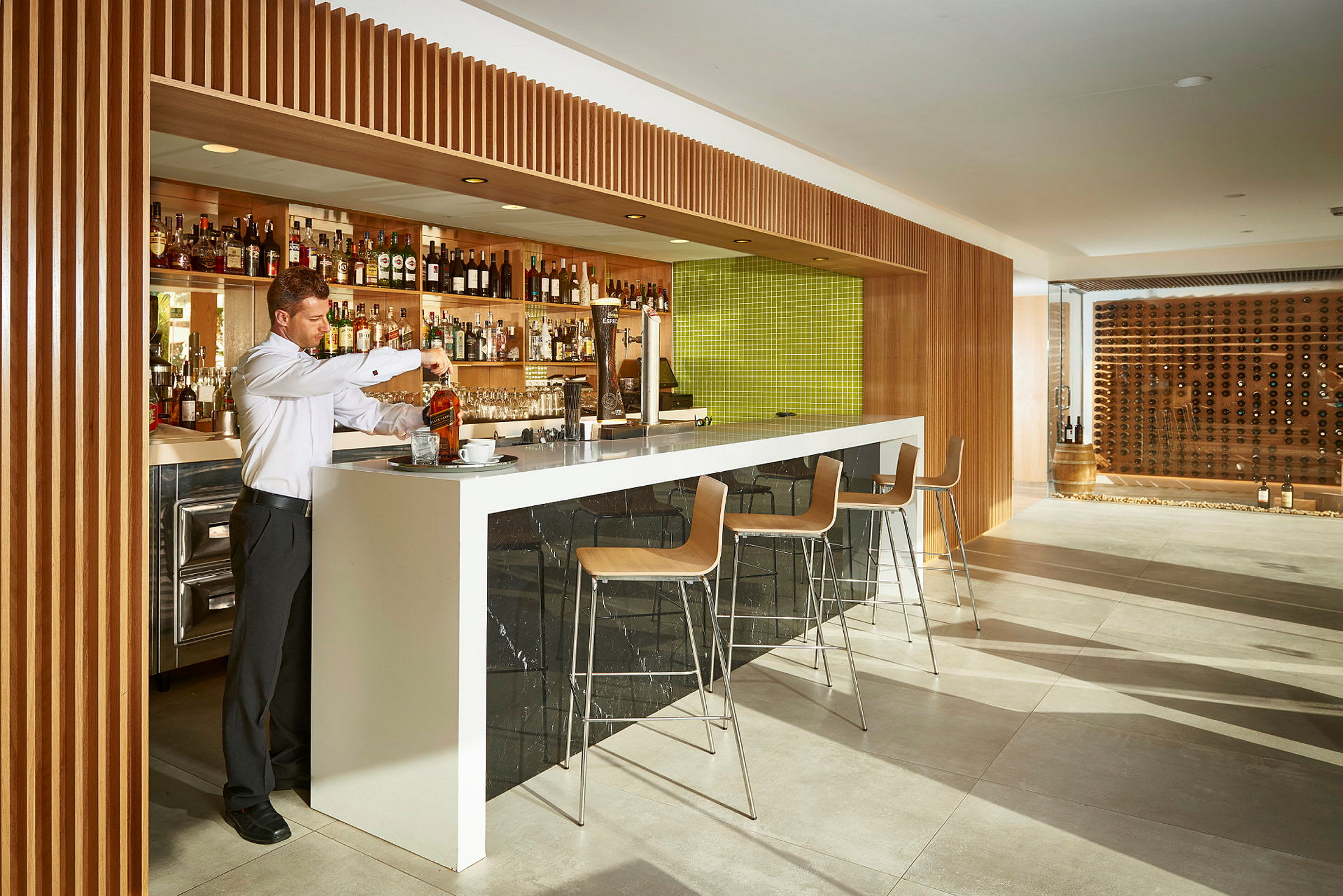 En bartender förbereder en drink vid en modern bar med träpaneler och en grön kaklad bakgrund. Barstolar väntar vid disken. (AI-genererad text)