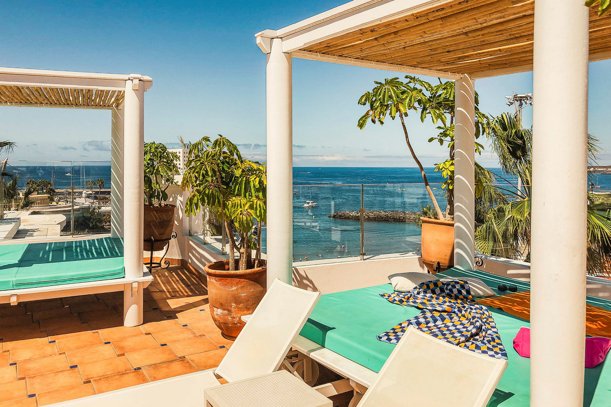 En solig terrass med utsikt över havet. En dagbädd med turkos madrass och färgglada tyger står under en pergola. Runt terrassen finns krukväxter och palmer. (AI-genererad text)