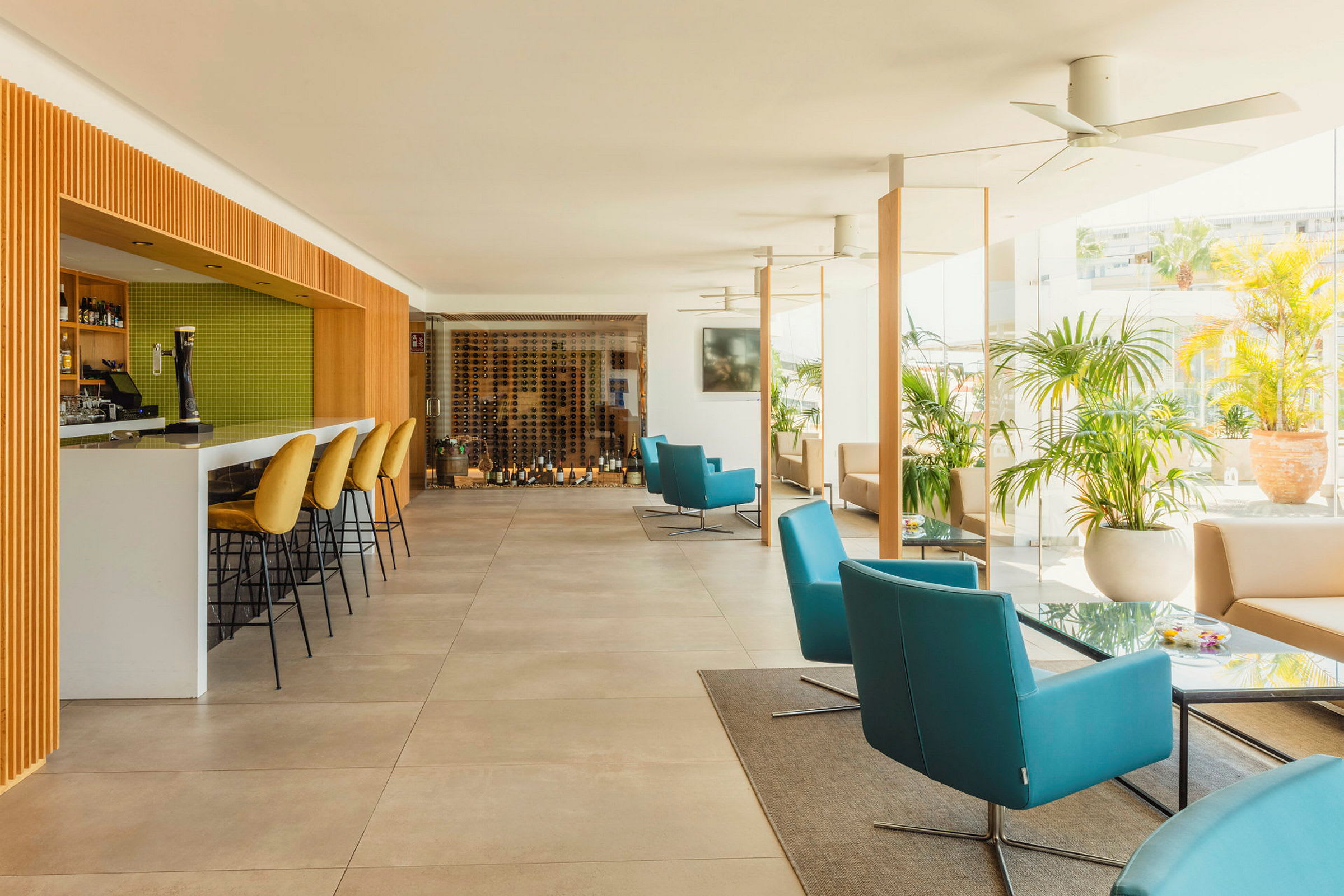 Ett modernt inrett caféområde med en bardisk och gröna kakelväggar, ryggstolar med guldfärgad klädsel, samt bekväma sittplatser med blå och beige fåtöljer. Grönska och glasväggar syns i bakgrunden. (AI-genererad text)