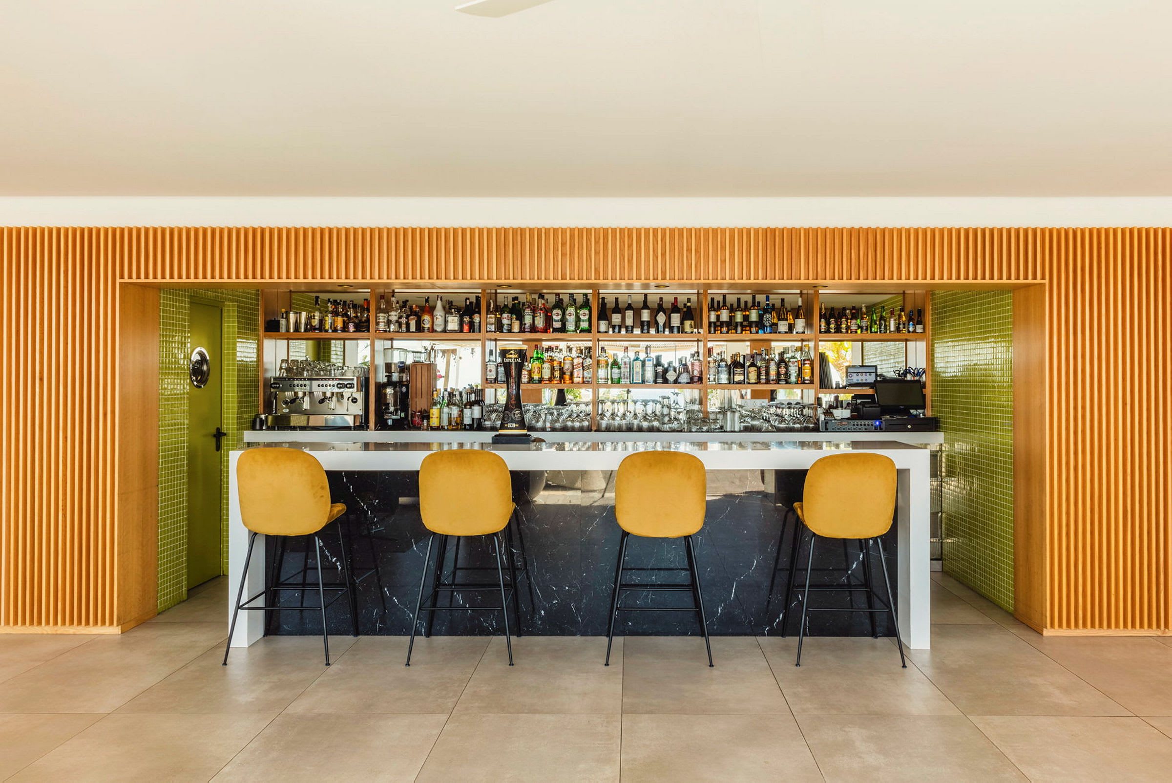 Ett interiörfoto av en modern bar med fyra guldfärgade barstolar framför en bardisk, och bakom detta syns hyllor fyllda med flaskor. (AI-genererad text)