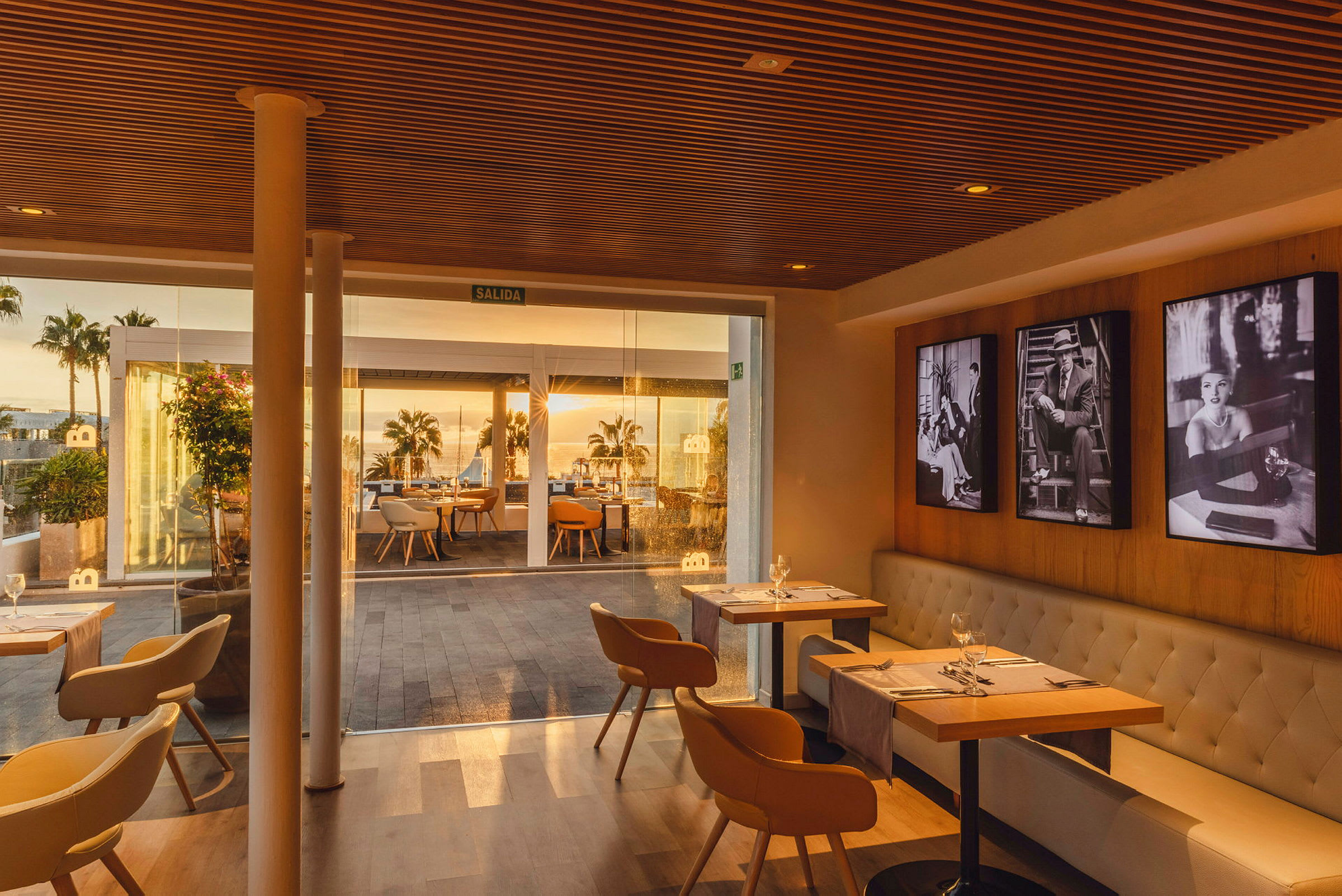 En inomhusdel av en restaurang med bord och stolar, samt glaspartier som visar utsikt mot en utomhusterrass med bord och palmer i solnedgång. (AI-genererad text)