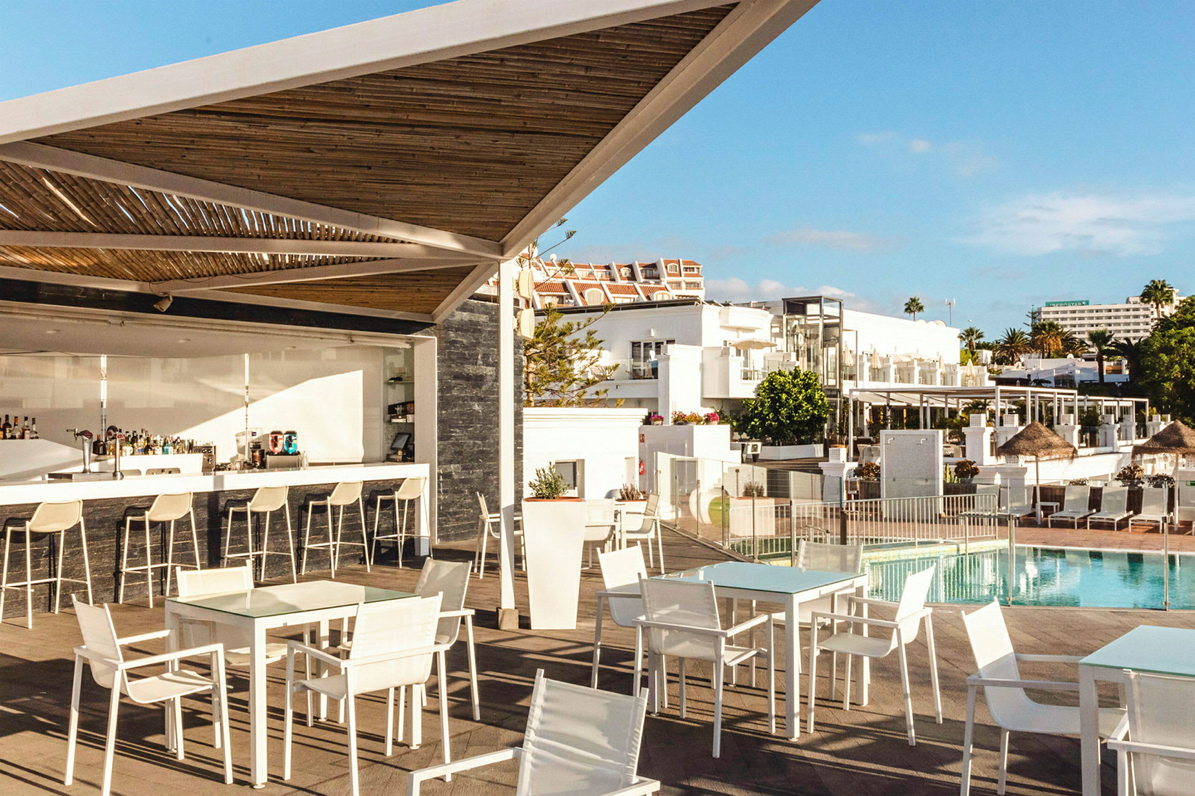 En utomhusbar med vita stolar och bord vid en pool och omgivning av vita byggnader. (AI-genererad text)