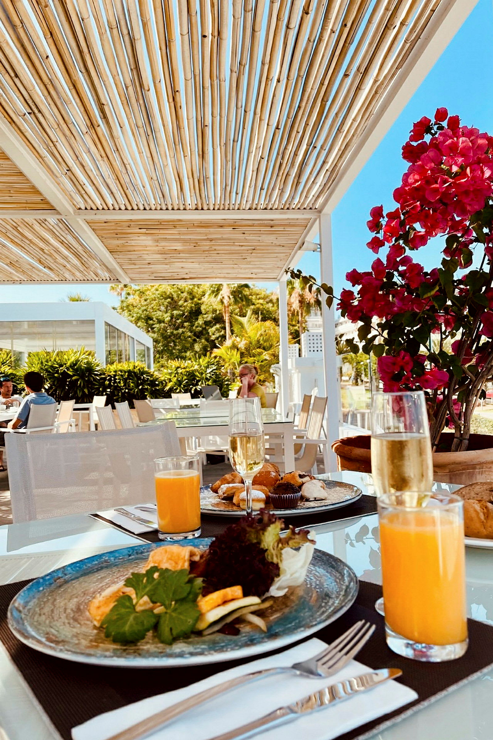 Ett bord är dukat med tallrikar, glas med juice och champagne, samt bestick. I bakgrunden syns en pergola, växter och människor som sitter vid andra bord. (AI-genererad text)