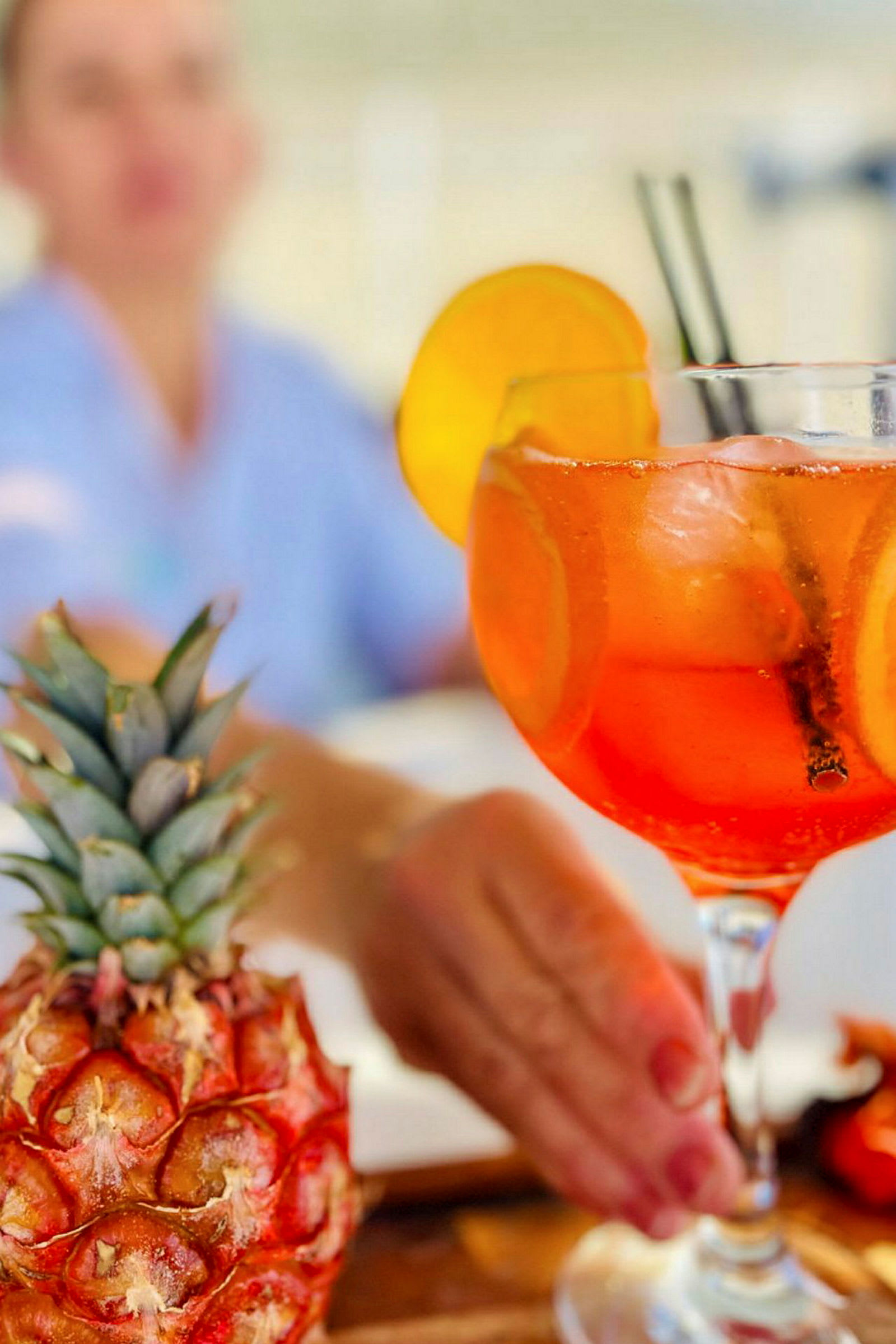 En närbild av en drink i ett glas med apelsinskivor och isbitar. En ananas syns i förgrunden. (AI-genererad text)