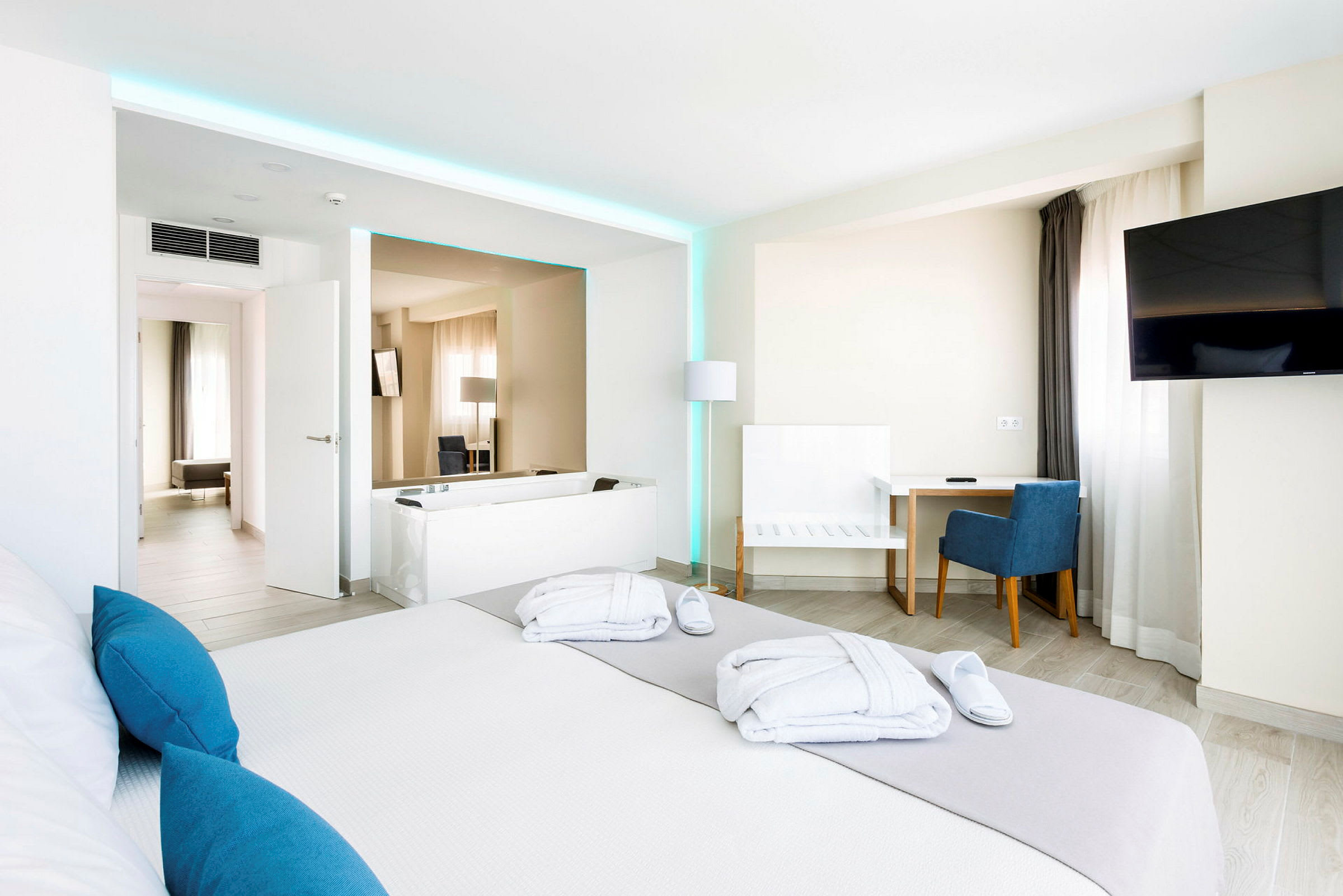 Ett modernt hotellrum med en stor säng, skrivbord och stol, en TV på väggen samt ett badkar. Ljusa väggar och inredning med blåa accenter. (AI-genererad text)