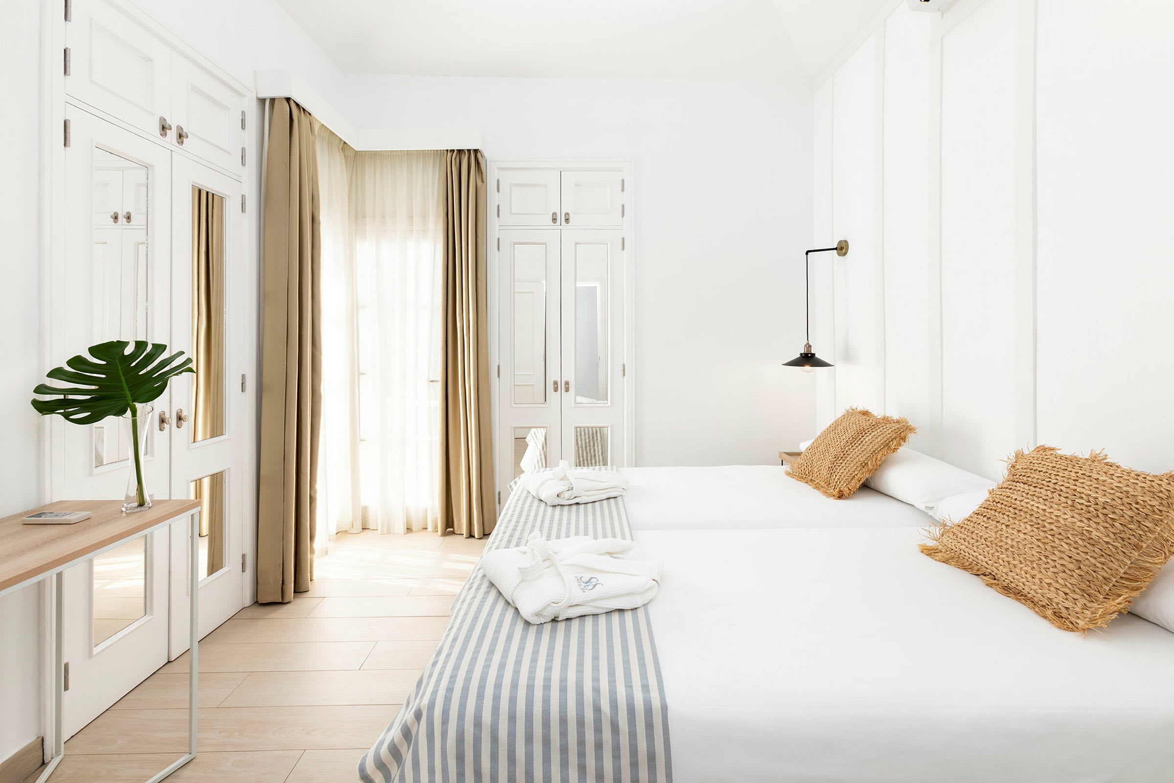 Ett ljust hotellrum med två enkelsängar, beige kuddar och ett bord med en grön växt i en glasvas. (AI-genererad text)