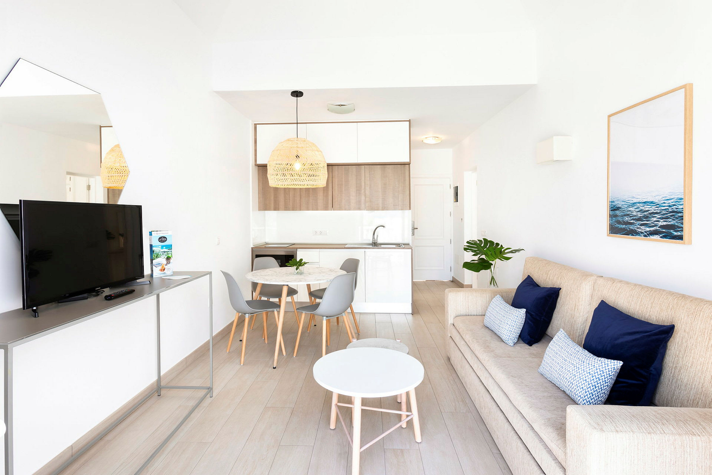 Ett modernt vardagsrum med en beige soffa, blå kuddar, en vit rund soffbord och en tv på ett stativ. Bredvid finns ett matområde med ett runt bord och grå stolar samt ett kök i ljusa färger. (AI-genererad text)
