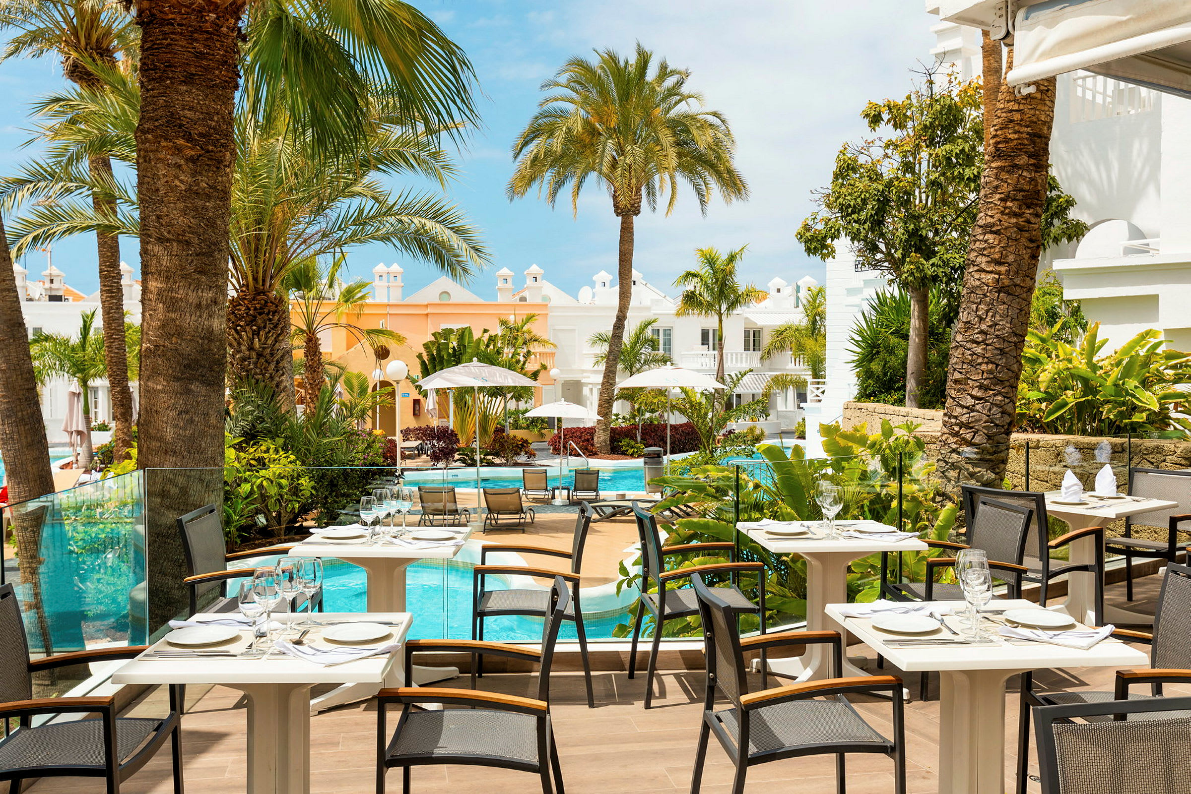 Bild av en restaurangterrass med utsikt över en pool omgiven av palmer och byggnader i ljusa färger. (AI-genererad text)