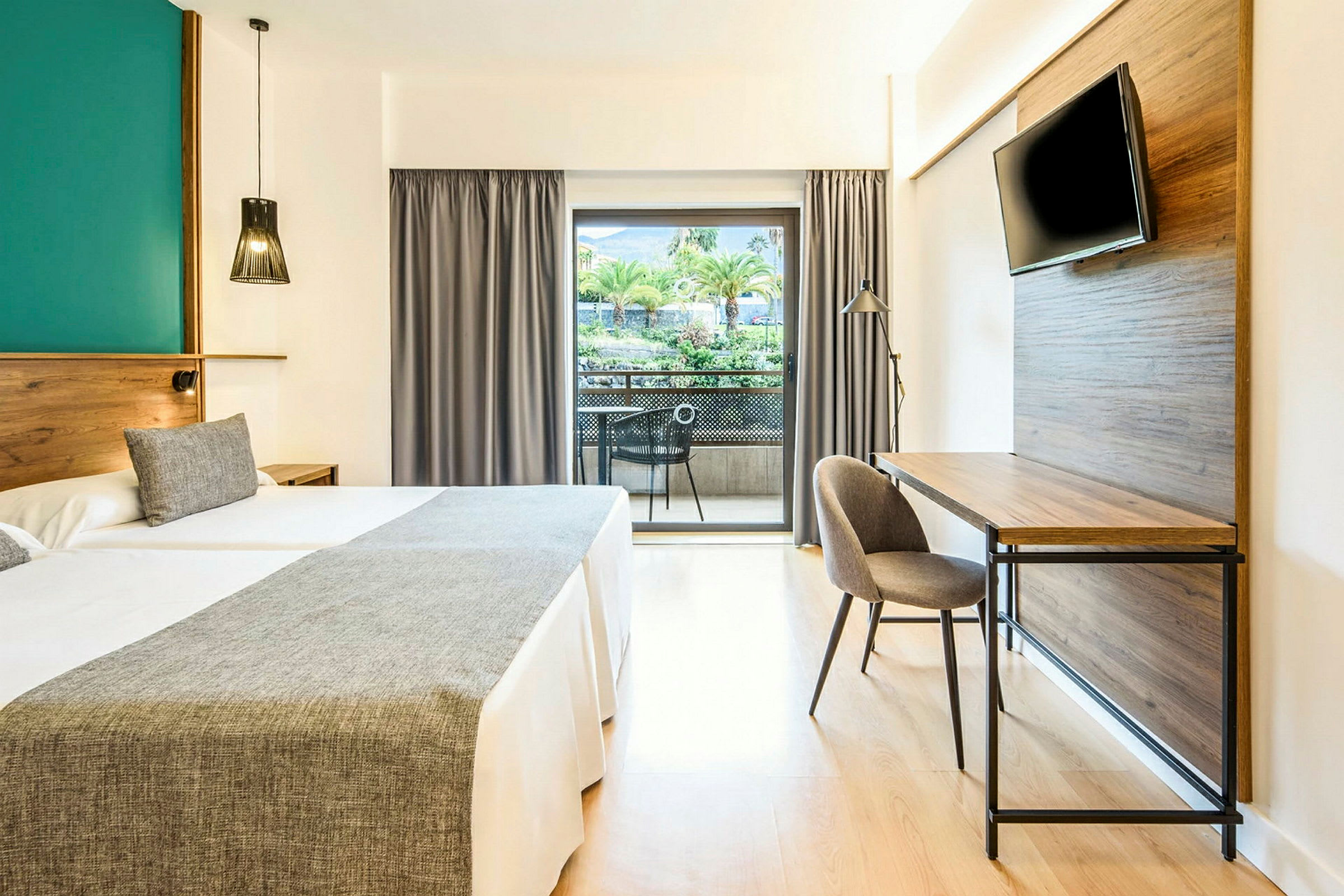 Hotellrum med två sängar, skrivbord, TV och balkong med utsikt över träd och grönska. (AI-genererad text)