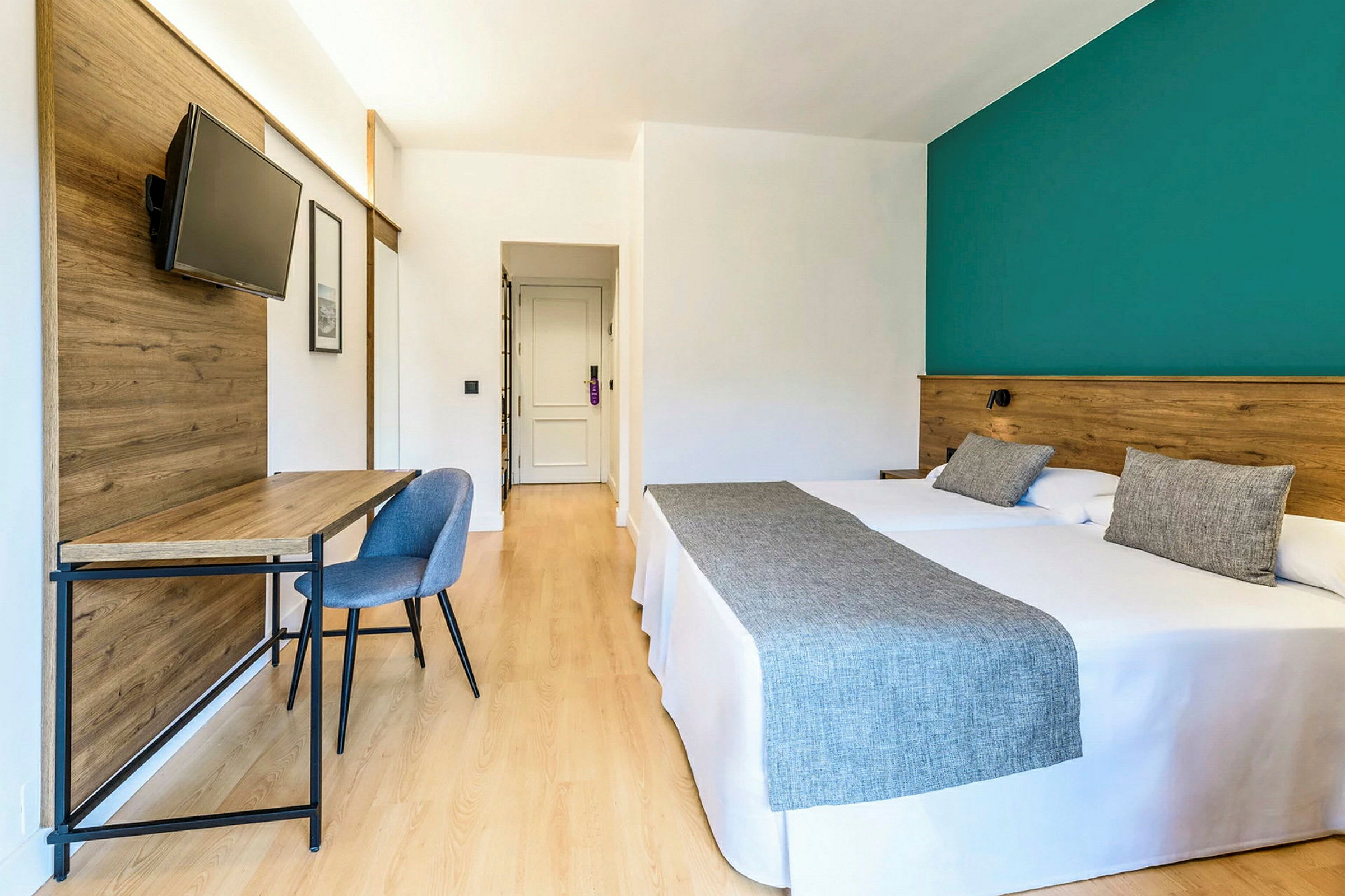 Ett hotellrum med två sängar, ett skrivbord med stol och en platt-TV på en träpanel. (AI-genererad text)