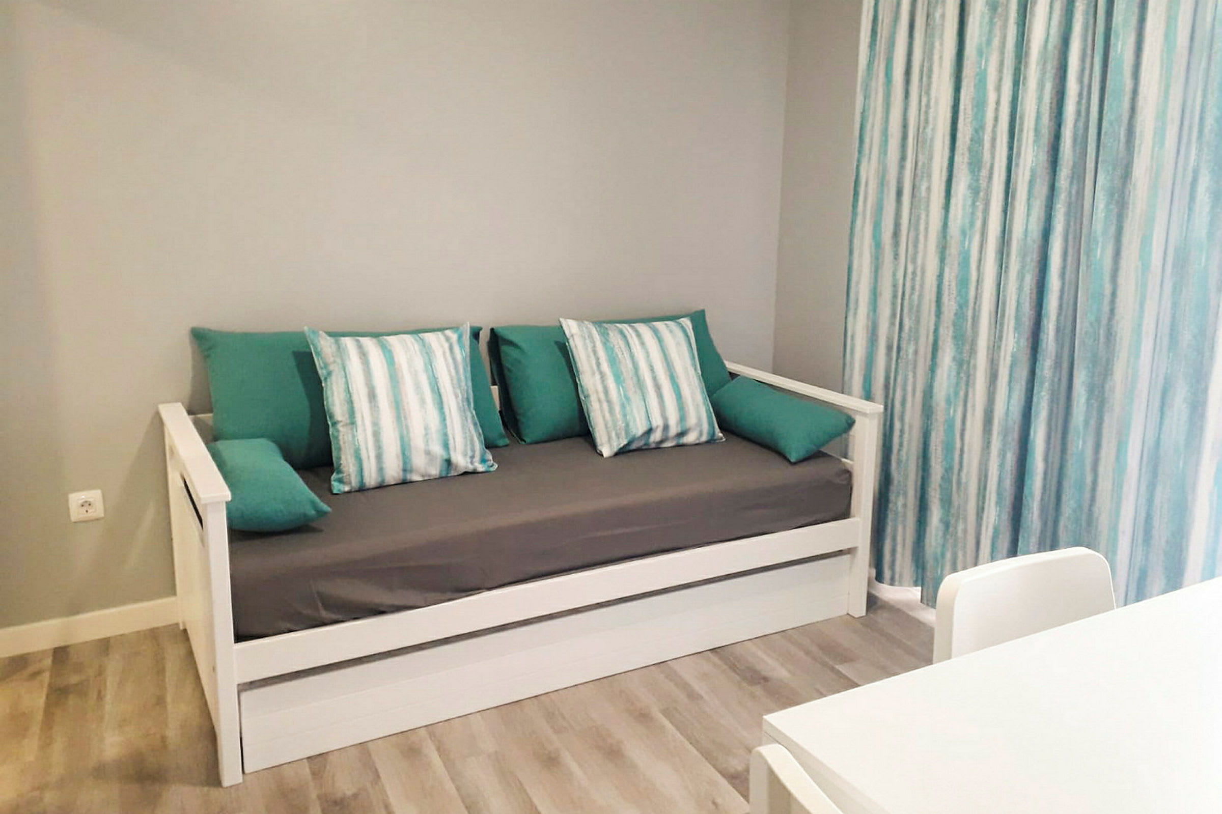 En träsoffa med grå klädsel och turkosa och randiga kuddar, bredvid ljusa gardiner. (AI-genererad text)