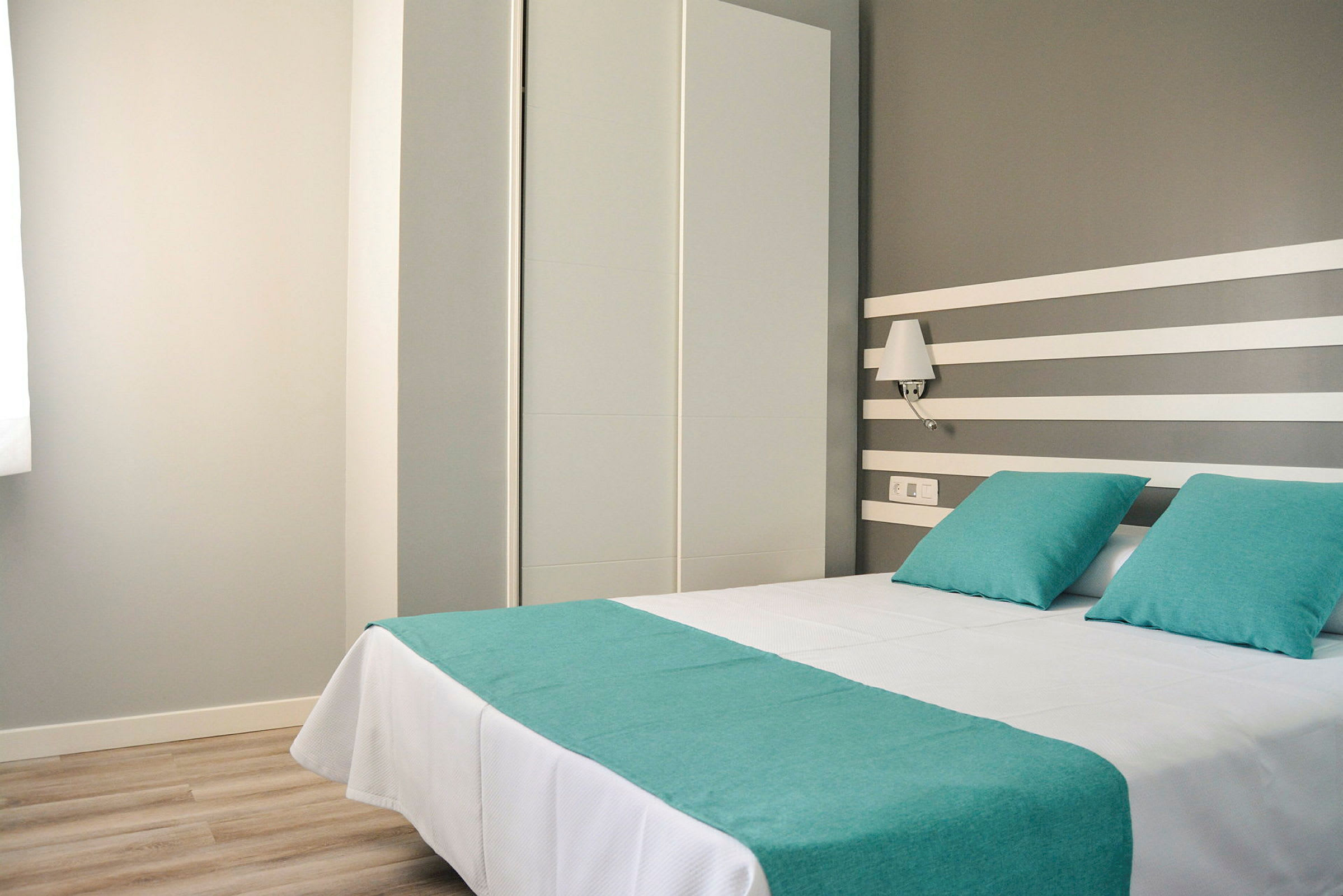 Hotellrum med en dubbelsäng bäddad med vita lakan och turkosa kuddar samt överkast. (AI-genererad text)