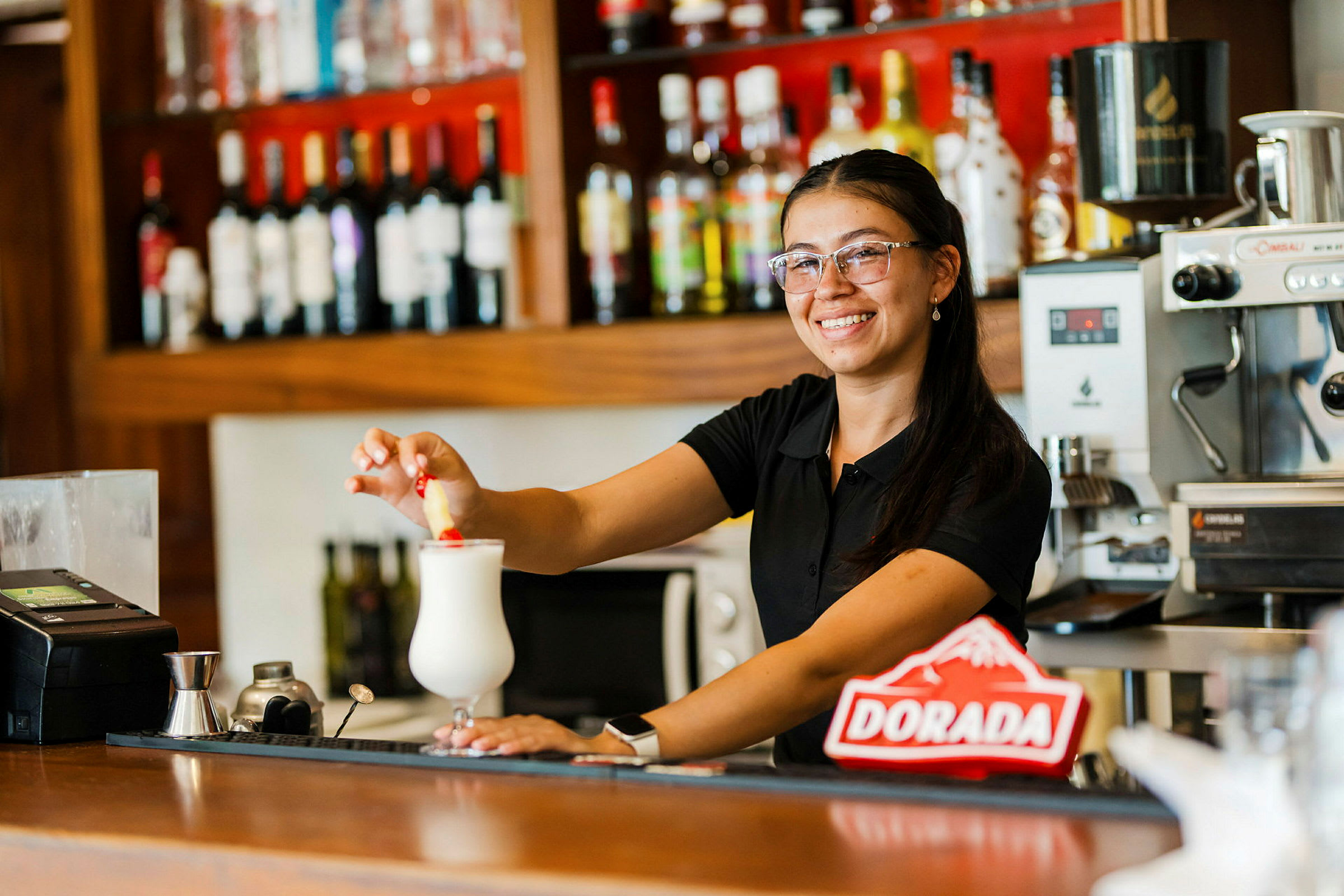 En person står vid en bardisk och dekorerar en vit drink med ett rött föremål. (AI-genererad text)