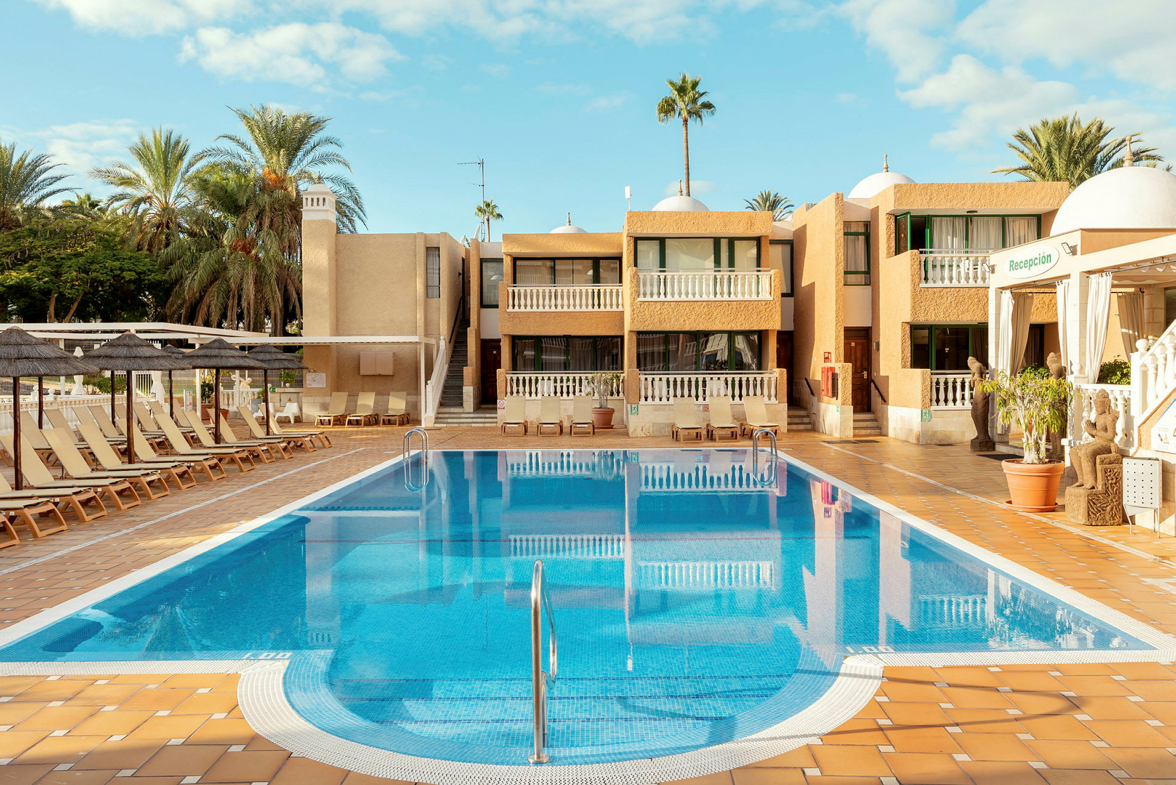 En hotellpool med solstolar och en byggnad i bakgrunden. (AI-genererad text)