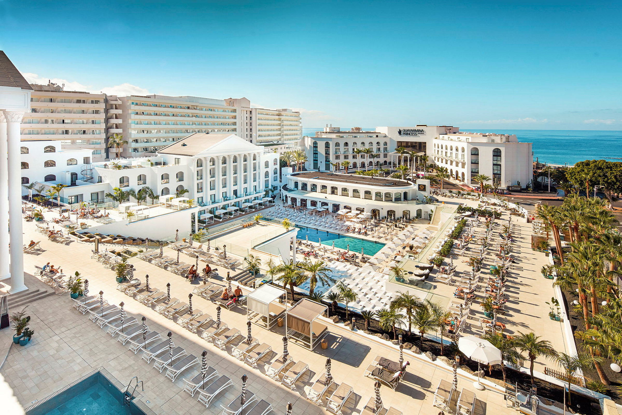 Utsikt över ett hotellområde med pooler, solstolar och vit arkitektur nära havet. (AI-genererad text)