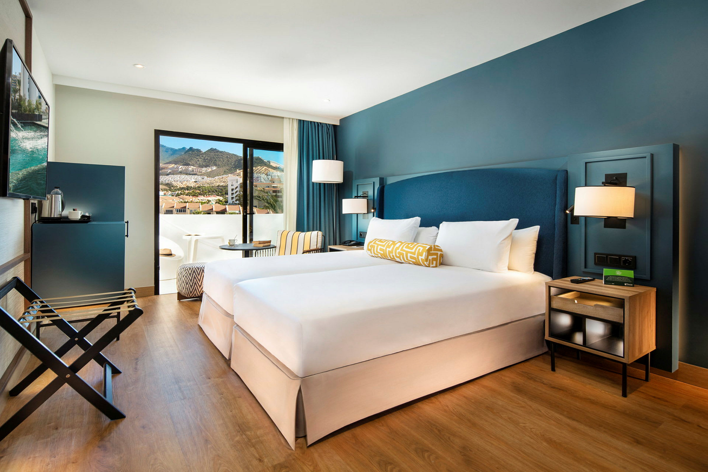 Ett hotellrum med två enkelsängar, blå väggar och en balkong med utsikt över bergen. (AI-genererad text)