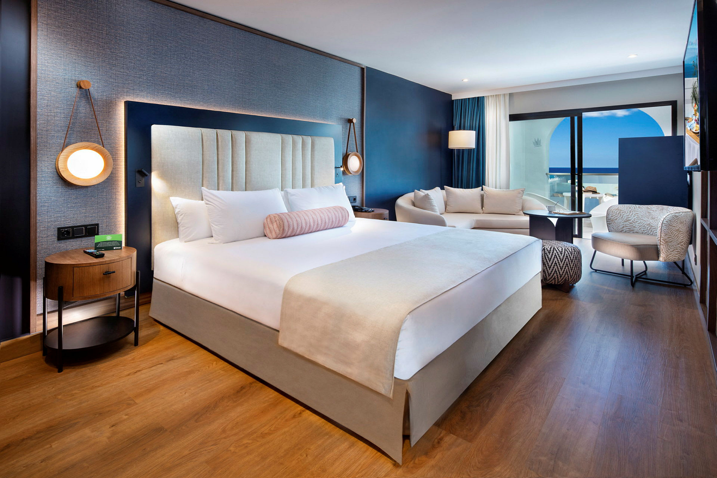 Ett modernt hotellrum med en stor säng, soffa och sittområde samt utsikt mot en terrass och havet. (AI-genererad text)