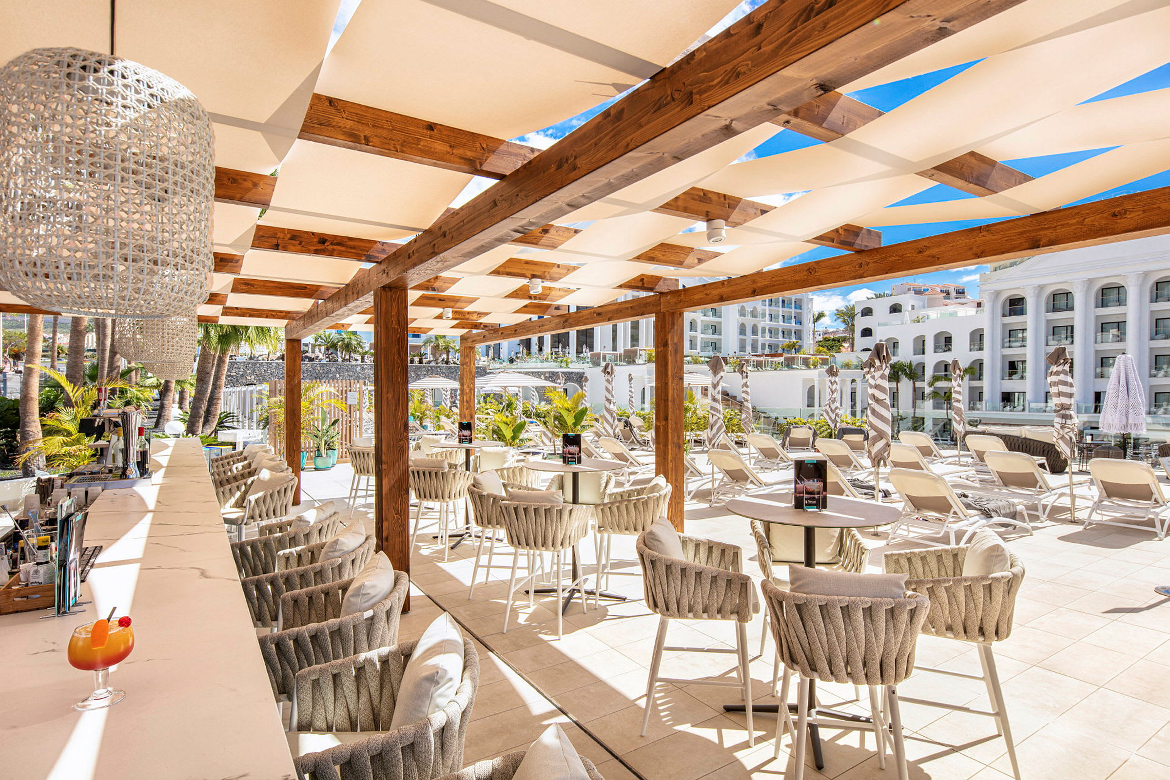 En utomhusbar med stolar och bord under en pergola, omgiven av solstolar och grönska. (AI-genererad text)