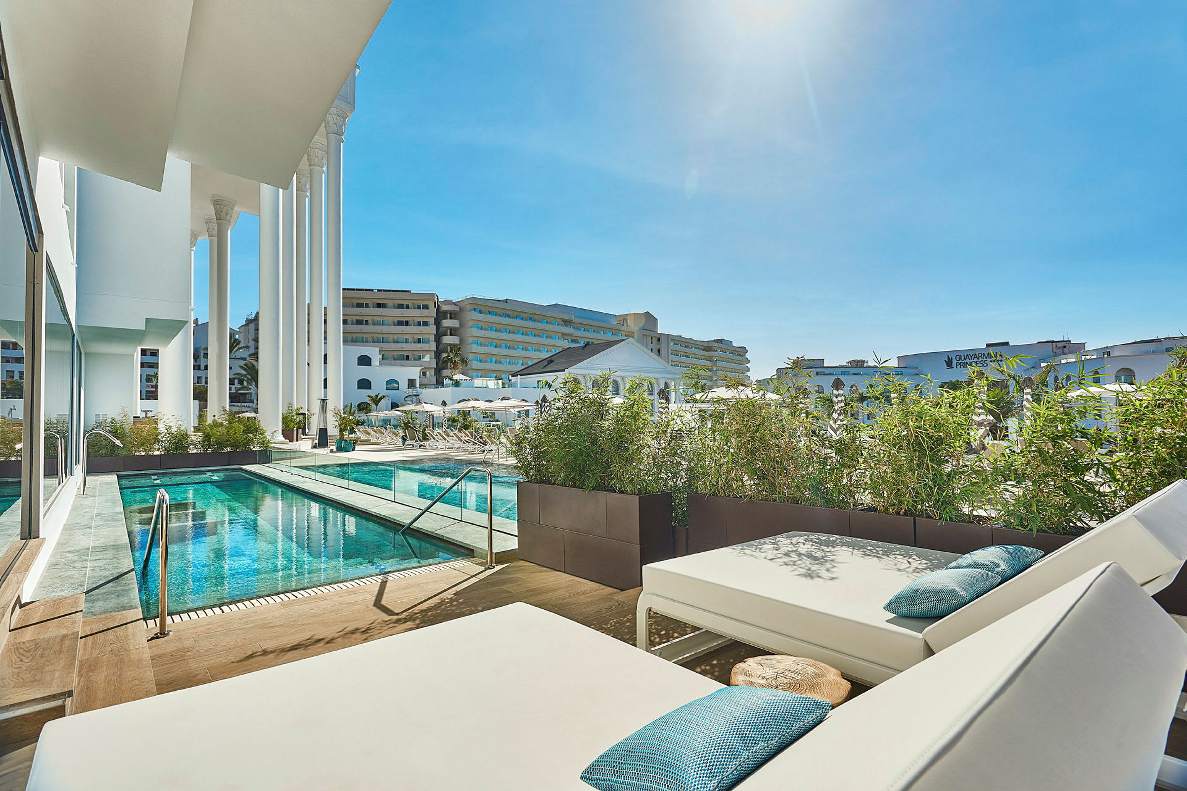 Utsikt över en pool och solstolar med hotellbyggnader i bakgrunden, omgivna av växter. (AI-genererad text)