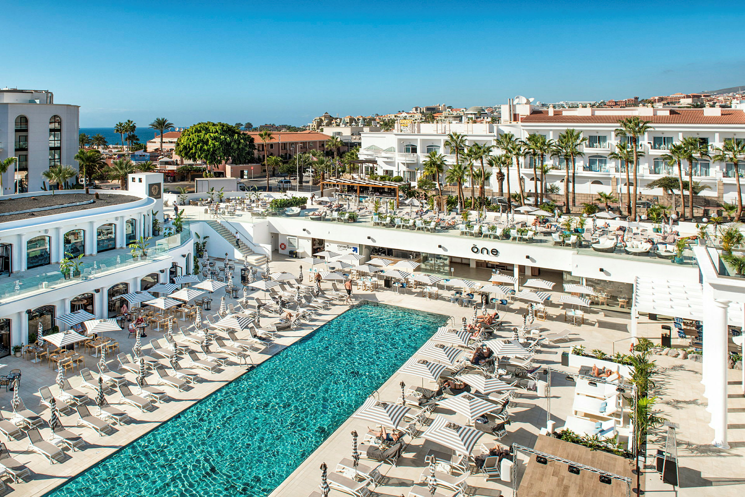 Bild av ett hotellområde med en utomhuspool, solstolar och parasoller, omgiven av byggnader och palmer. (AI-genererad text)