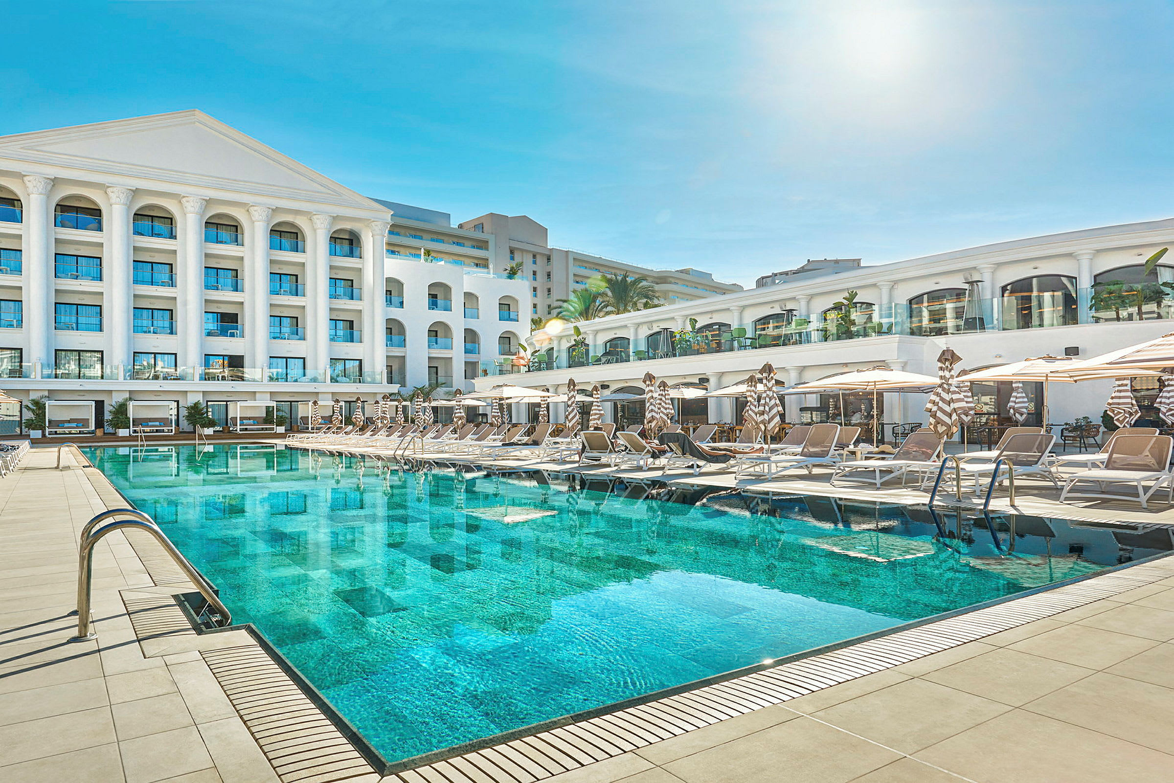 En hotellbyggnad med en stor swimmingpool och solstolar framför. (AI-genererad text)