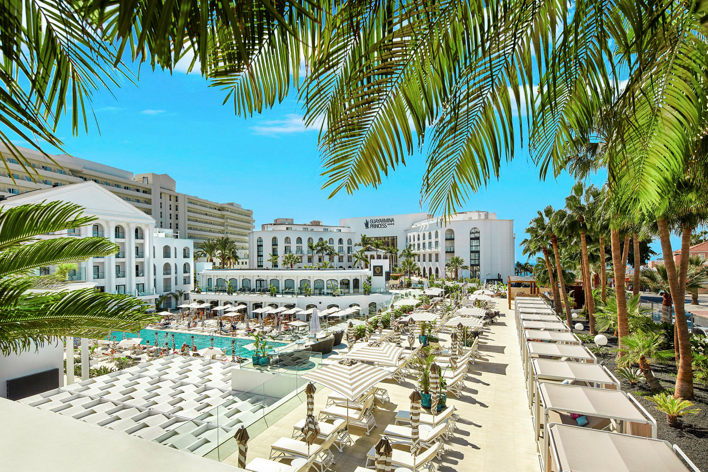 Utsikt över ett hotellkomplex med en pool och omgivande solstolar, inramat av palmer. (AI-genererad text)