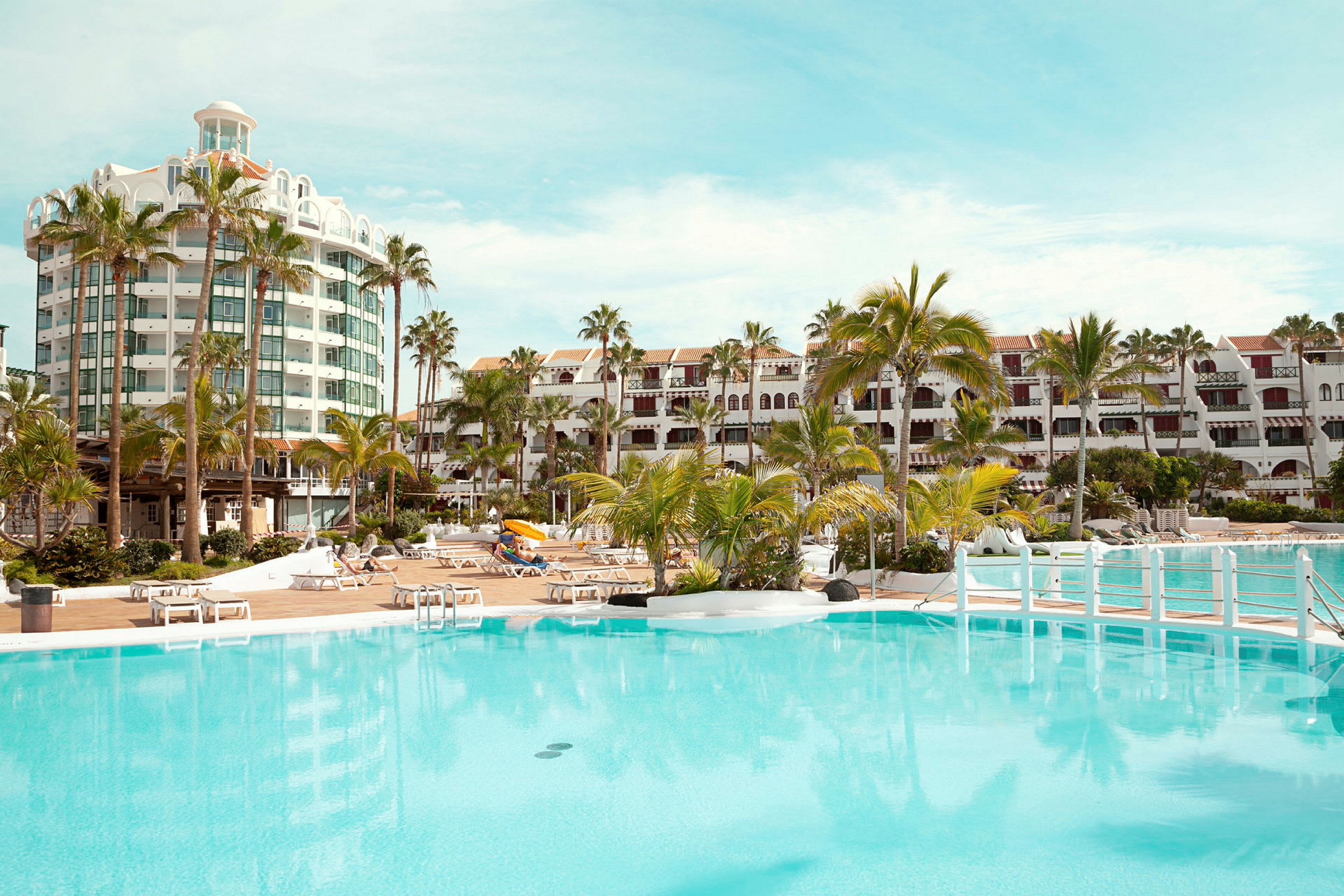 Utomhuspool med omgivande solstolar framför hotellbyggnader och palmer. (AI-genererad text)