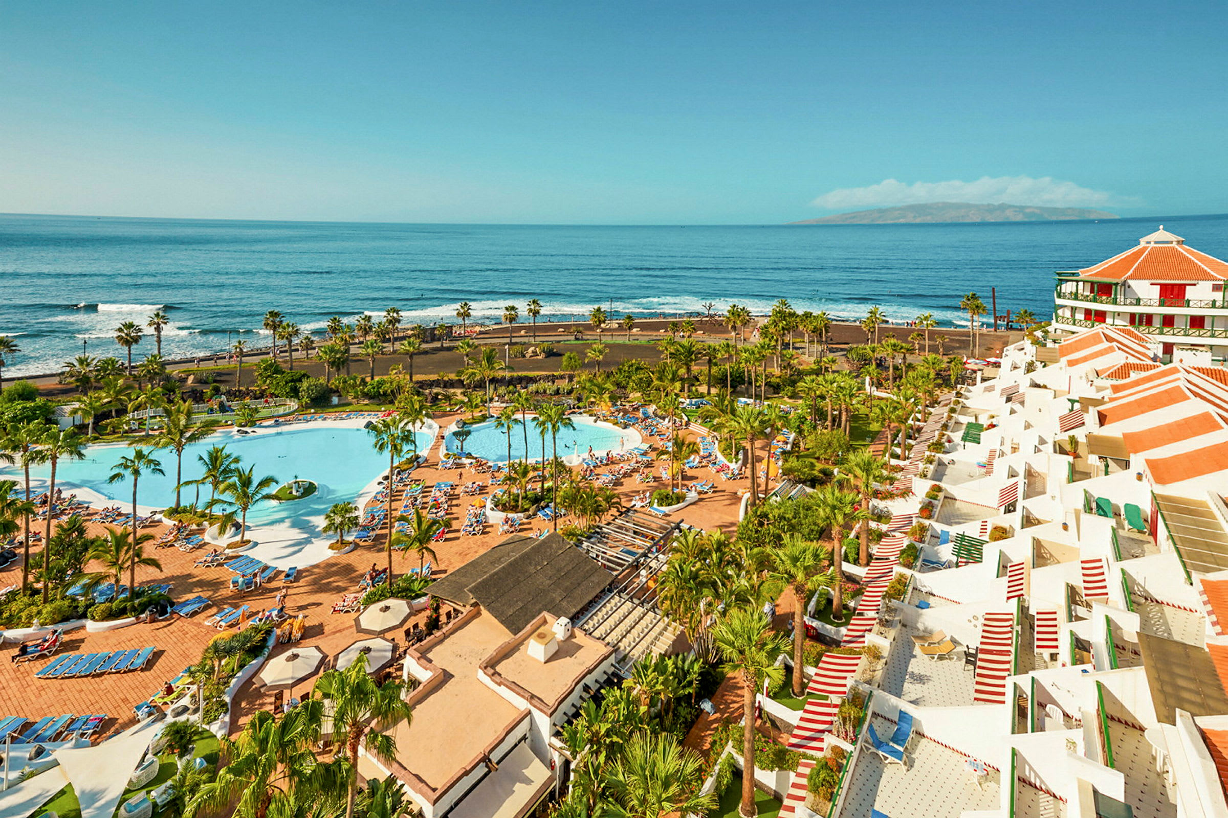 Vy över ett semesterhotell med poolområde, palmer och havet i bakgrunden. (AI-genererad text)