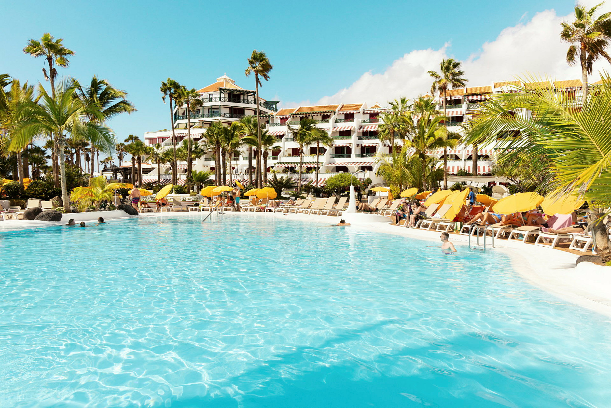 Stor pool omgiven av solstolar och parasoller framför hotellbyggnad. (AI-genererad text)