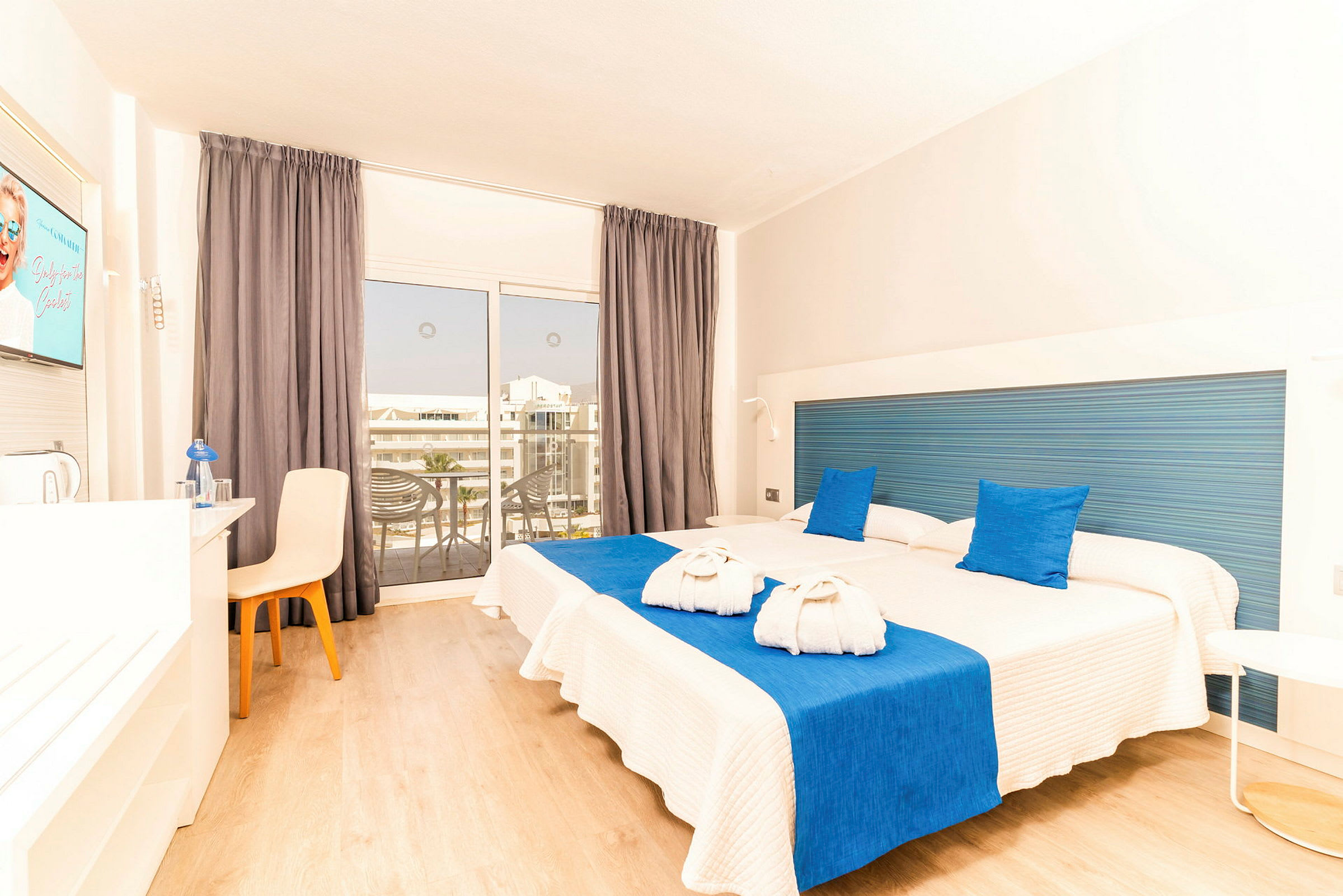 Ett hotellrum med två enkelsängar täckta med vita överkast och blå detaljer, en TV på väggen och en balkong med utsikt. (AI-genererad text)