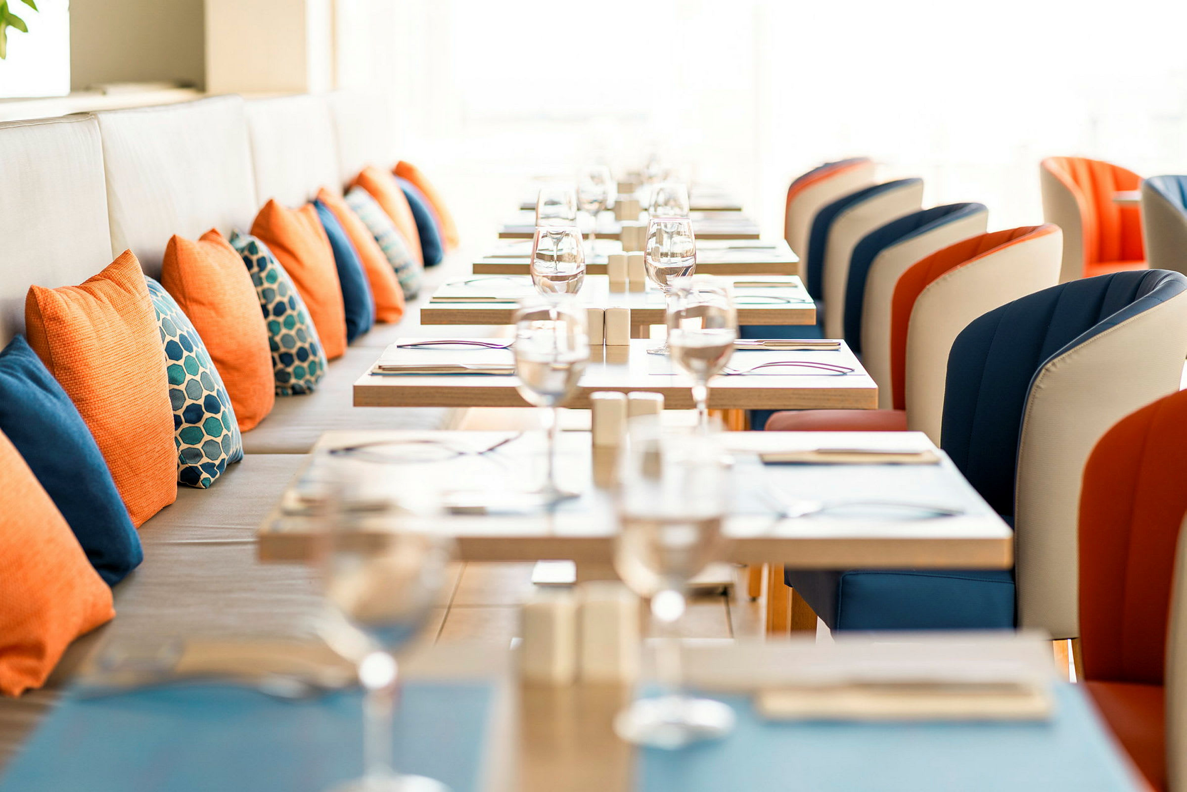 På bilden syns en dukad restaurang med bord och stolar. Stolarna är i orange och blå färger, och orangea och blå kuddar pryder sittplatser mot väggen. (AI-genererad text)