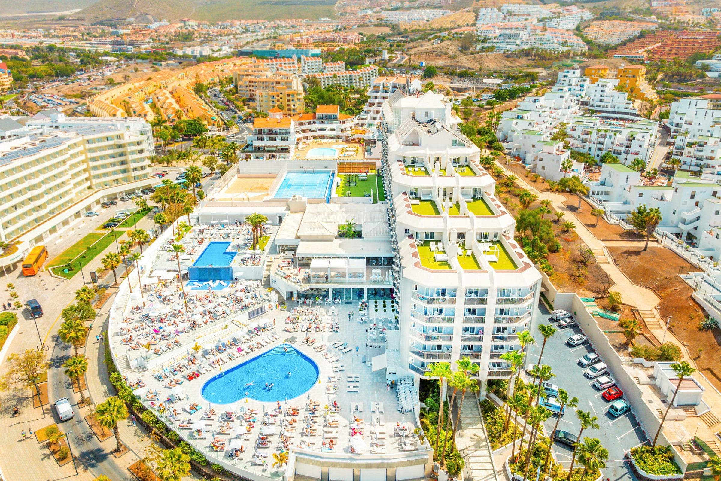 En flygfoto av ett hotellområde med pooler, solstolar och omgivande byggnader. (AI-genererad text)