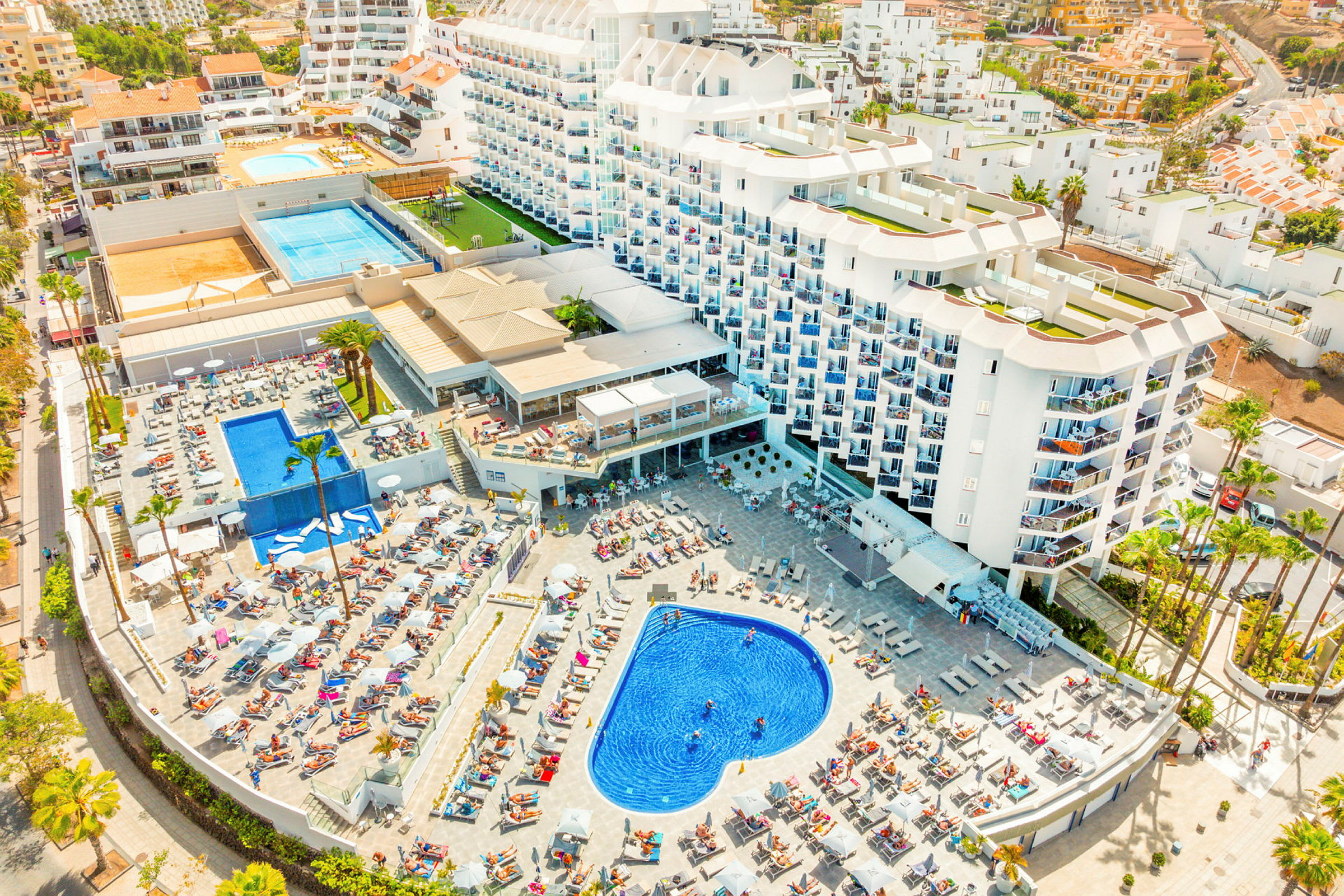 Hotellkomplex med poolområde och solstolar, omgivet av byggnader och grönska. (AI-genererad text)