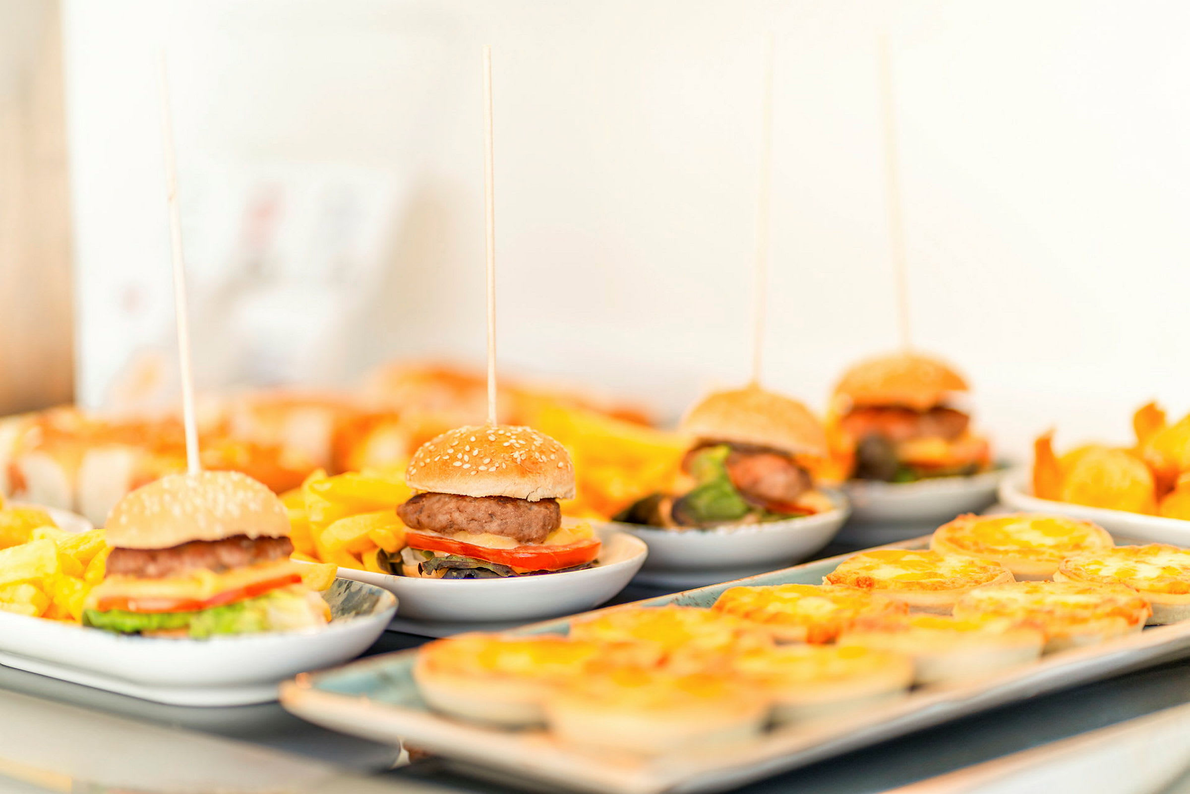 Små hamburgare på tallrikar med tillbehör och andra maträtter, uppställda på ett bord. (AI-genererad text)