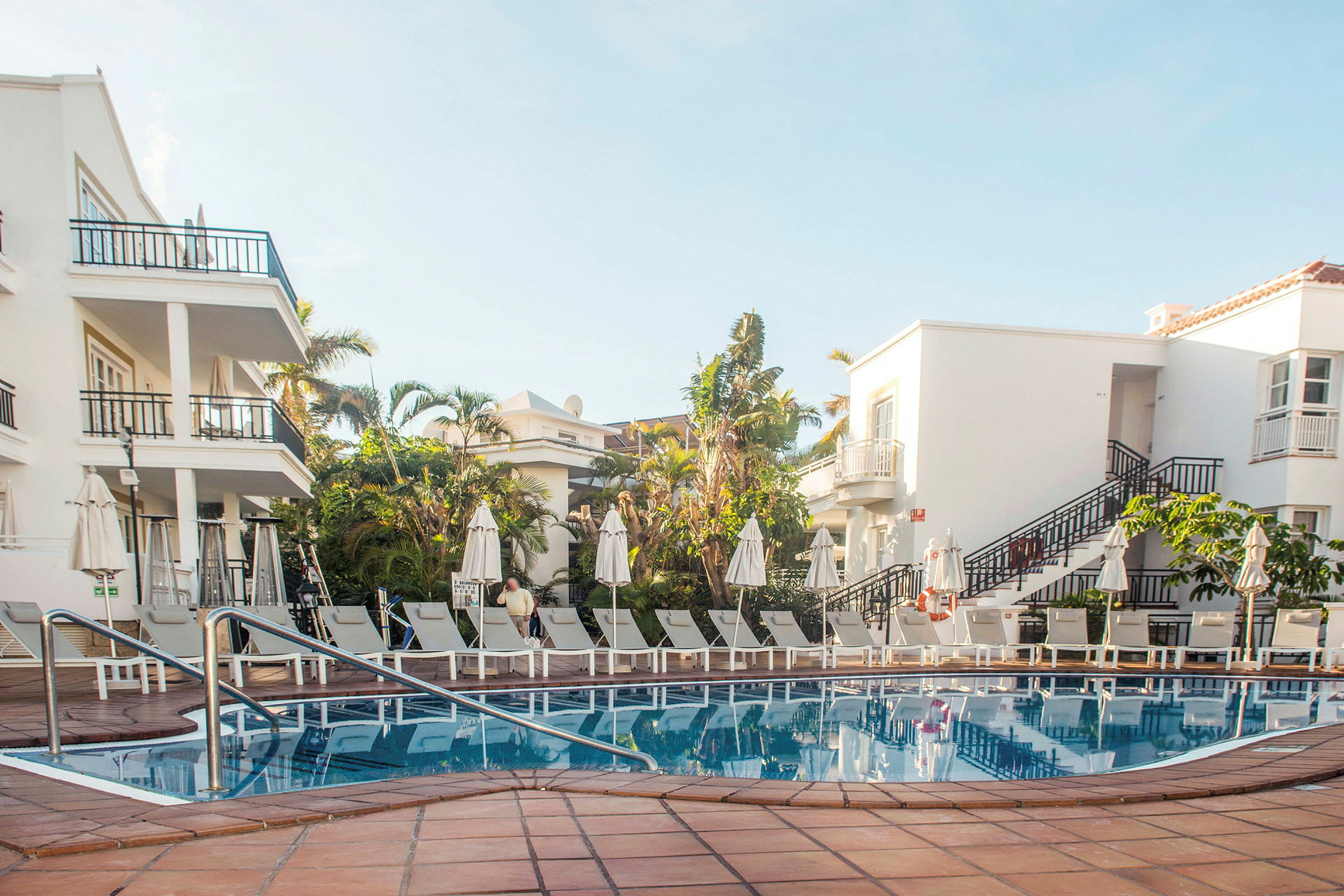 Uteområde med pool, solstolar och vita byggnader i bakgrunden. (AI-genererad text)