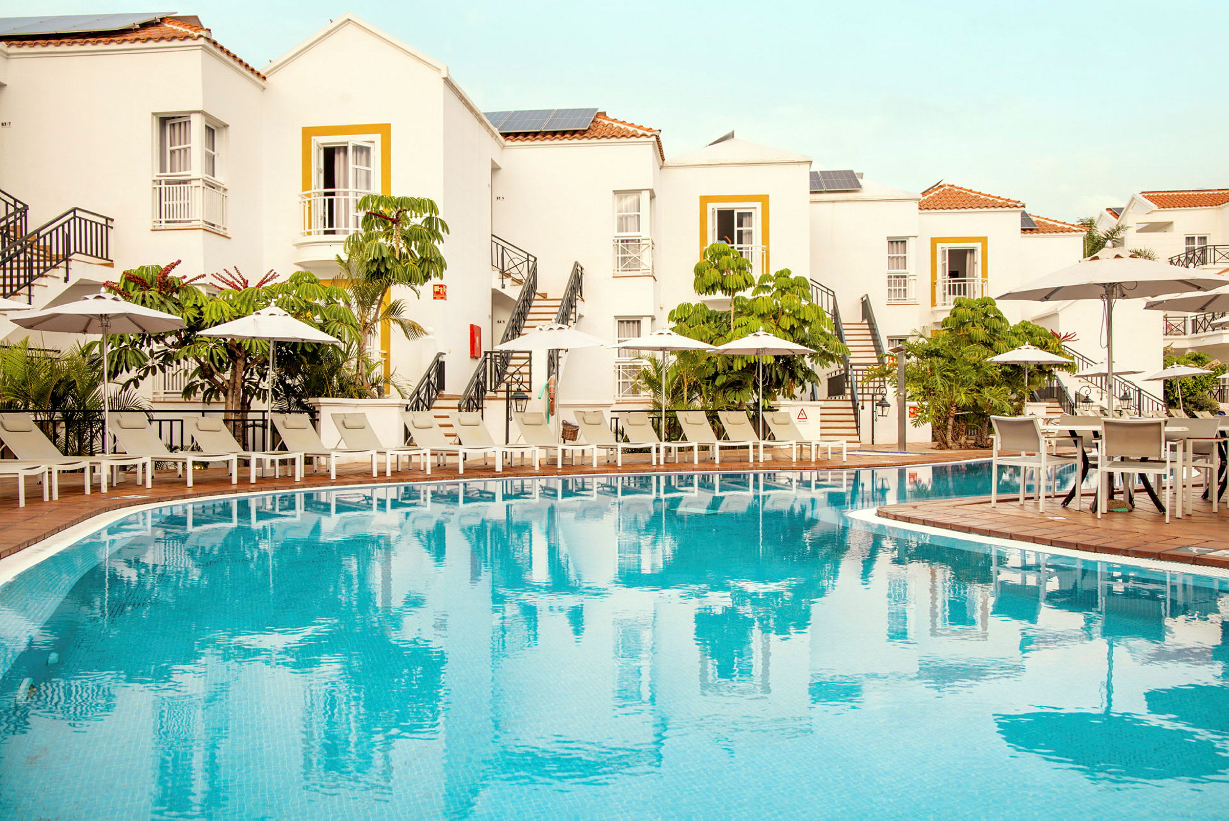 Hotellbyggnad med vita fasader och gul dörrkant, en pool framför, omgiven av solstolar och parasoller. (AI-genererad text)