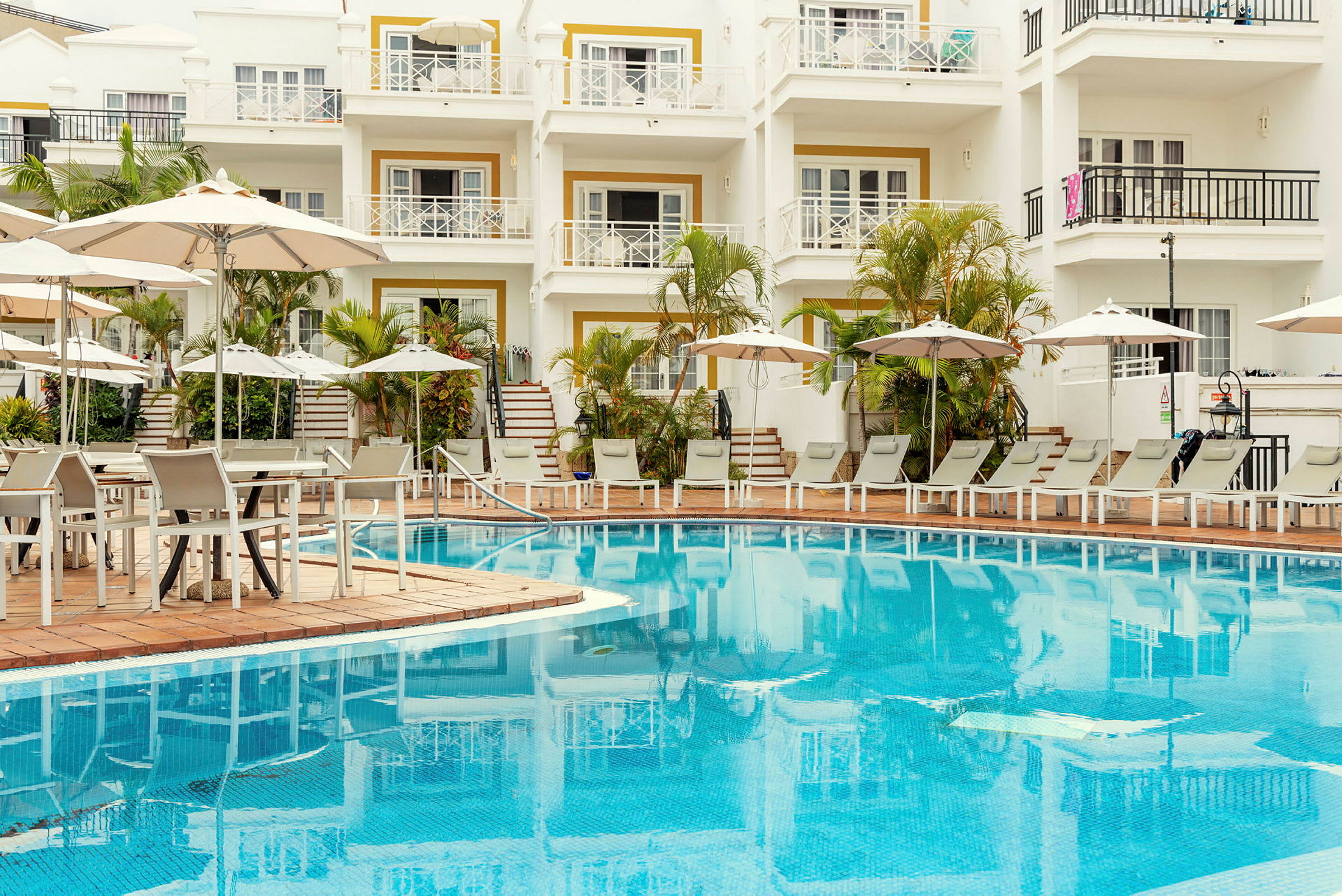 Bild av en hotellpool med solstolar, parasoller och ett flerbostadshus i bakgrunden. (AI-genererad text)