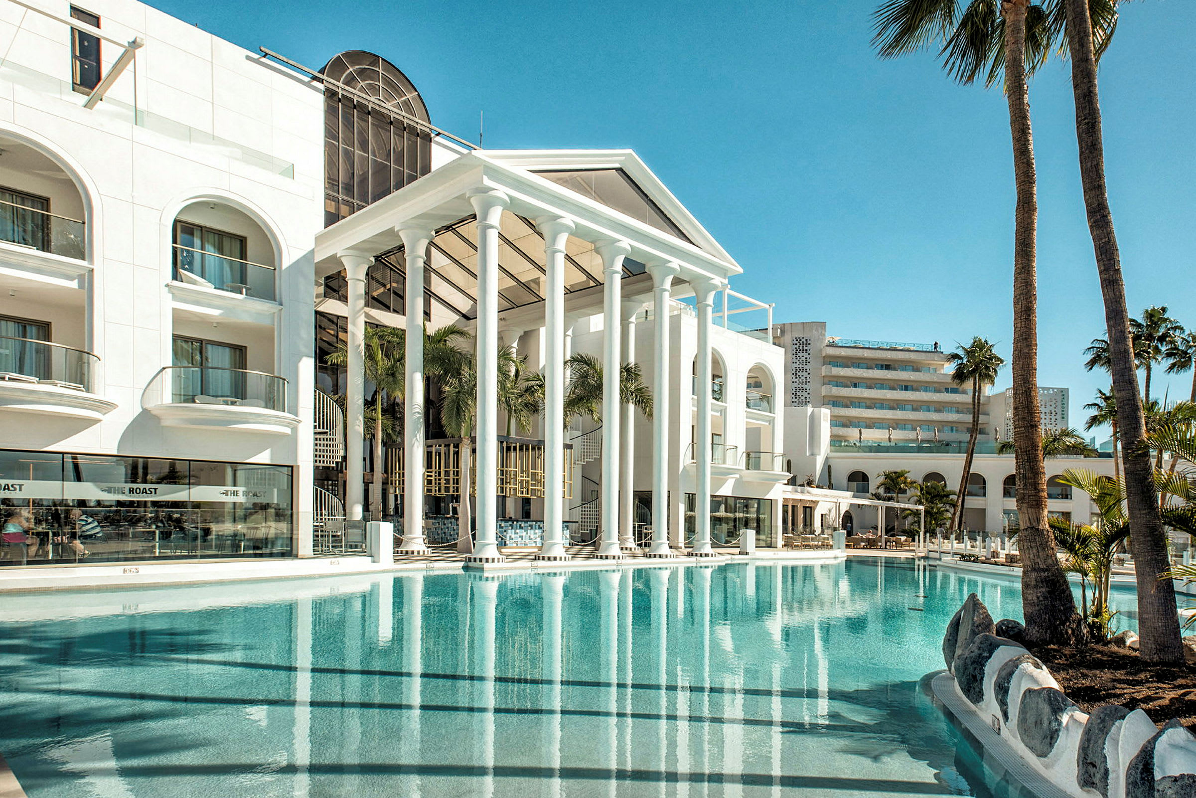 Hotellfasad med pool och palmer i förgrunden. (AI-genererad text)