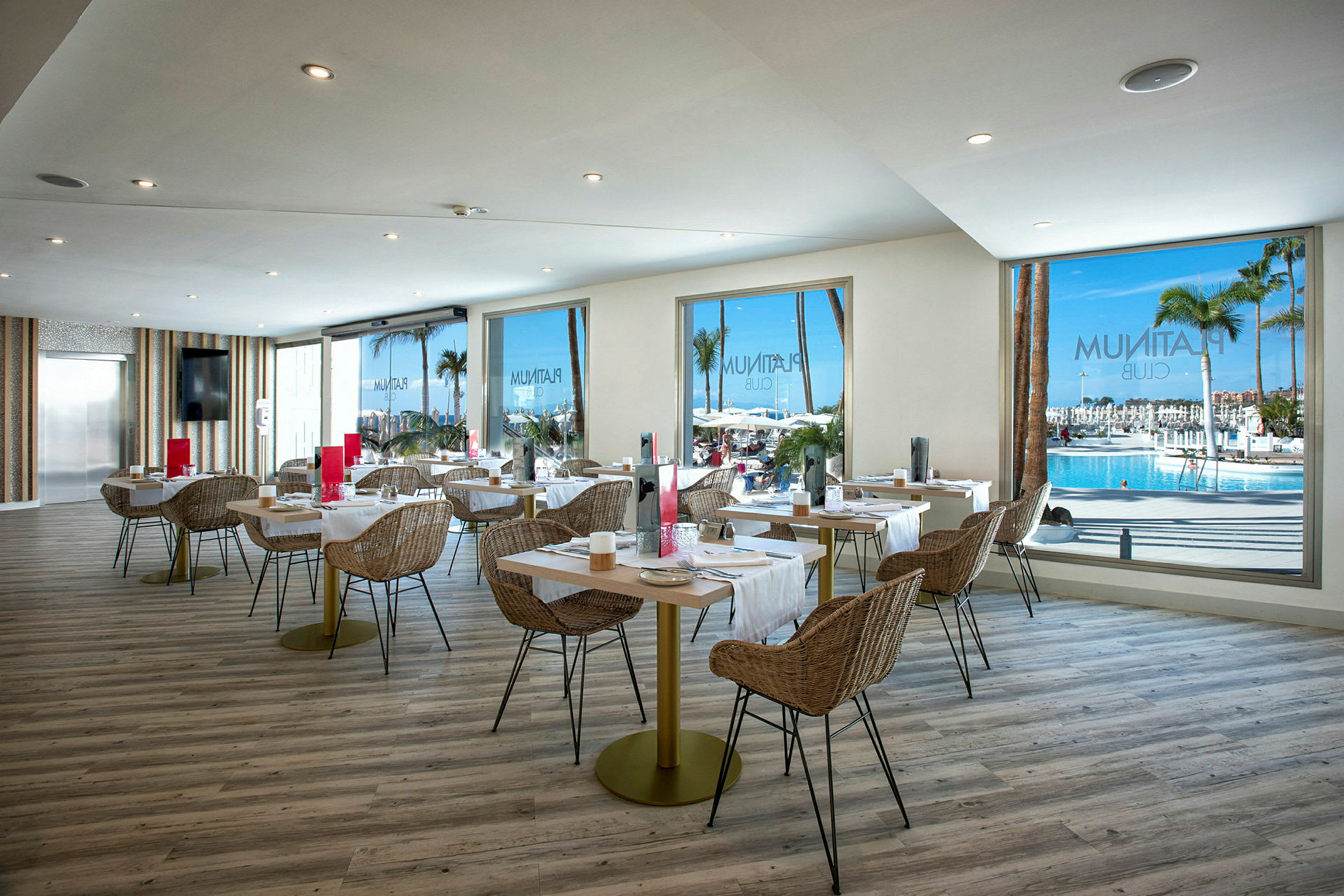 Inredning av en ljus restaurang med utsikt mot en pool och palmträd genom stora fönster. (AI-genererad text)