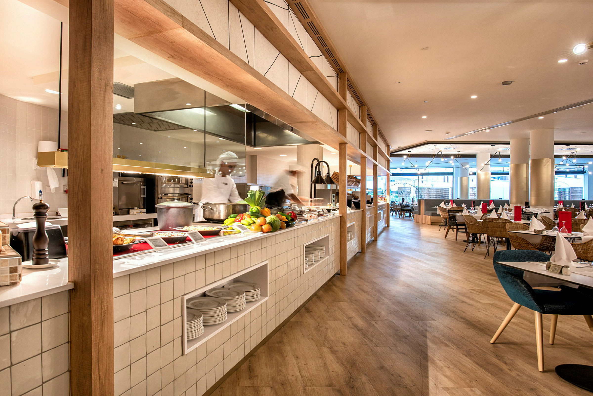 Bild av en modern restaurang med öppet kök, bänk med färska råvaror och matservering samt sittplatser bakom. (AI-genererad text)